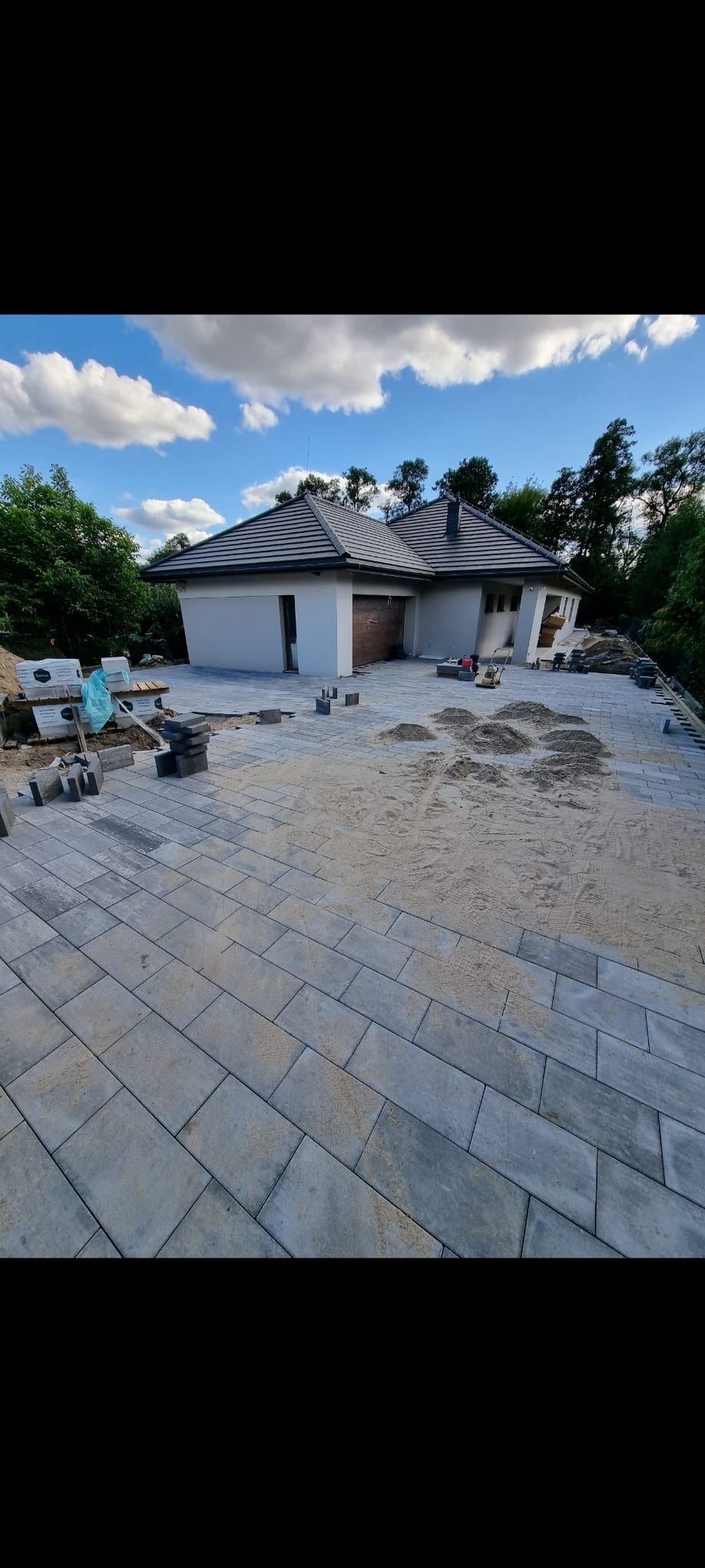 Układanie szarej kostki brukowej przed domem jednorodzinnym. Częściowo wyłożony podjazd z widocznym piaskiem i stertą kostki. Błękitne niebo z chmurami w tle.