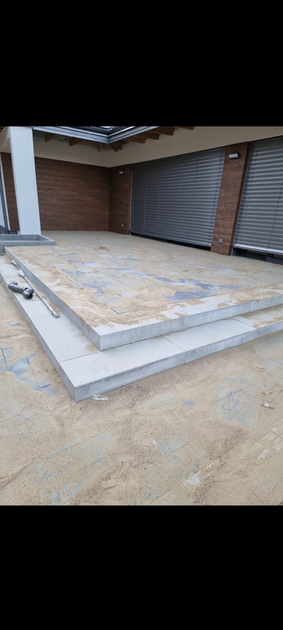 Ułożone schody z jasnoszarej kostki brukowej, zasypane piaskiem, prowadzące na taras. Widoczny fragment elewacji budynku z brązowymi panelami.