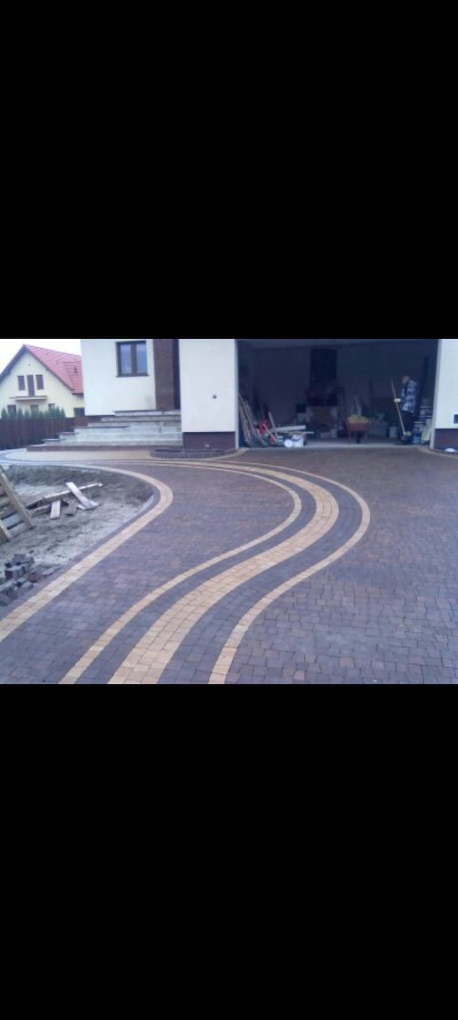 Układanie kostki brukowej w łukowate wzory na podjeździe. Częściowo ułożona kostka, widoczne narzędzia i materiały budowlane. Dom jednorodzinny w tle.