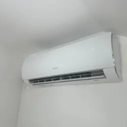 Biała klimatyzacja Sinclair, zamontowana na białej ścianie w rogu pomieszczenia. Widoczny wyświetlacz temperatury, żaluzje wlotu powietrza i obudowa urządzenia.