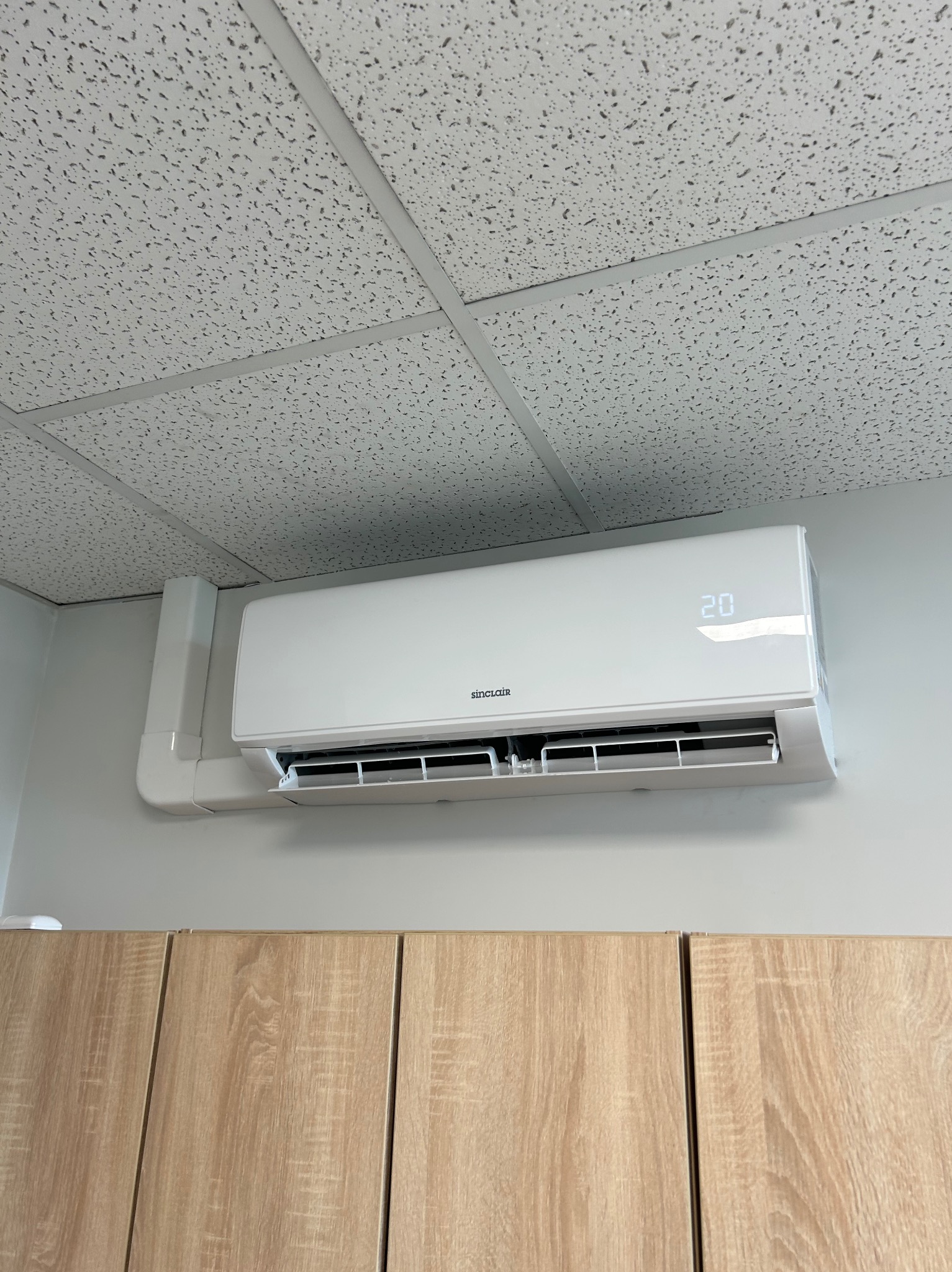 Biały klimatyzator Sinclair zamontowany na szarej ścianie nad drewnianymi szafkami. Widoczny fragment sufitu kasetonowego. Na wyświetlaczu urządzenia widoczna temperatura 20 stopni.