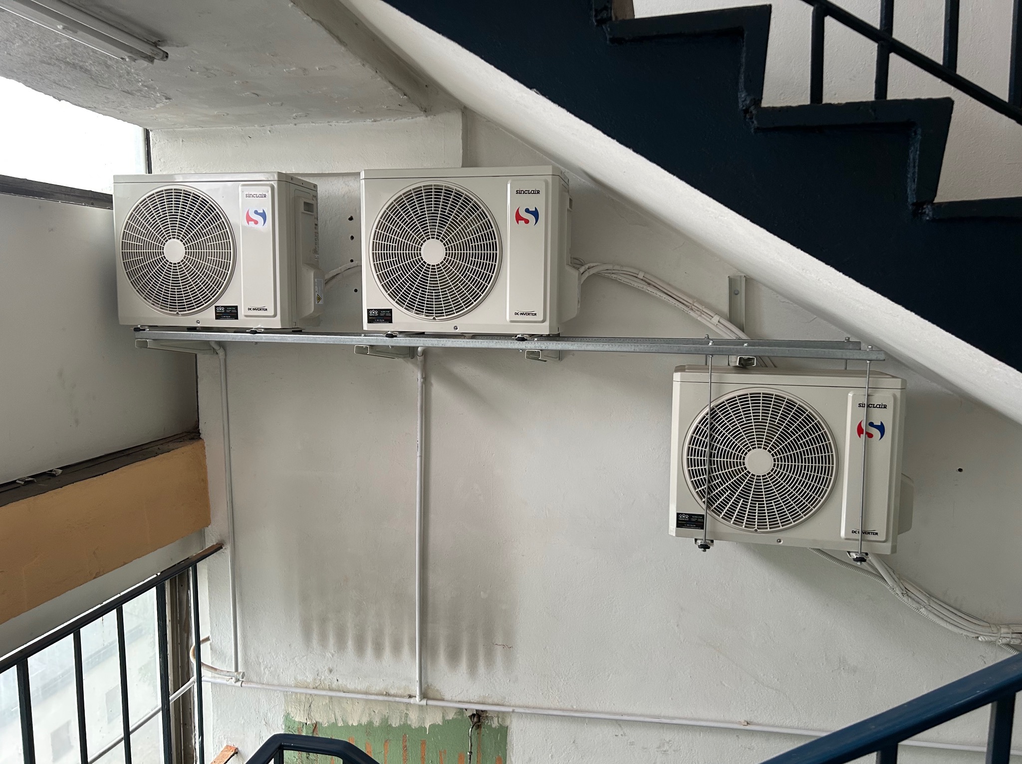 Trzy klimatyzatory Sinclair zamontowane na metalowej konstrukcji w klatce schodowej. Widoczne schody, okno i instalacja elektryczna.