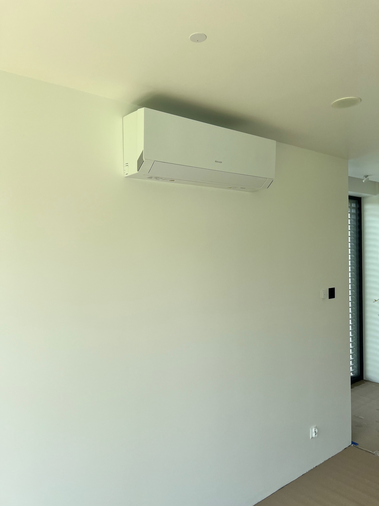 Nowoczesny, biały klimatyzator ścienny marki Sinclair zamontowany w jasnym, minimalistycznym wnętrzu z widocznymi głośnikami sufitowymi.