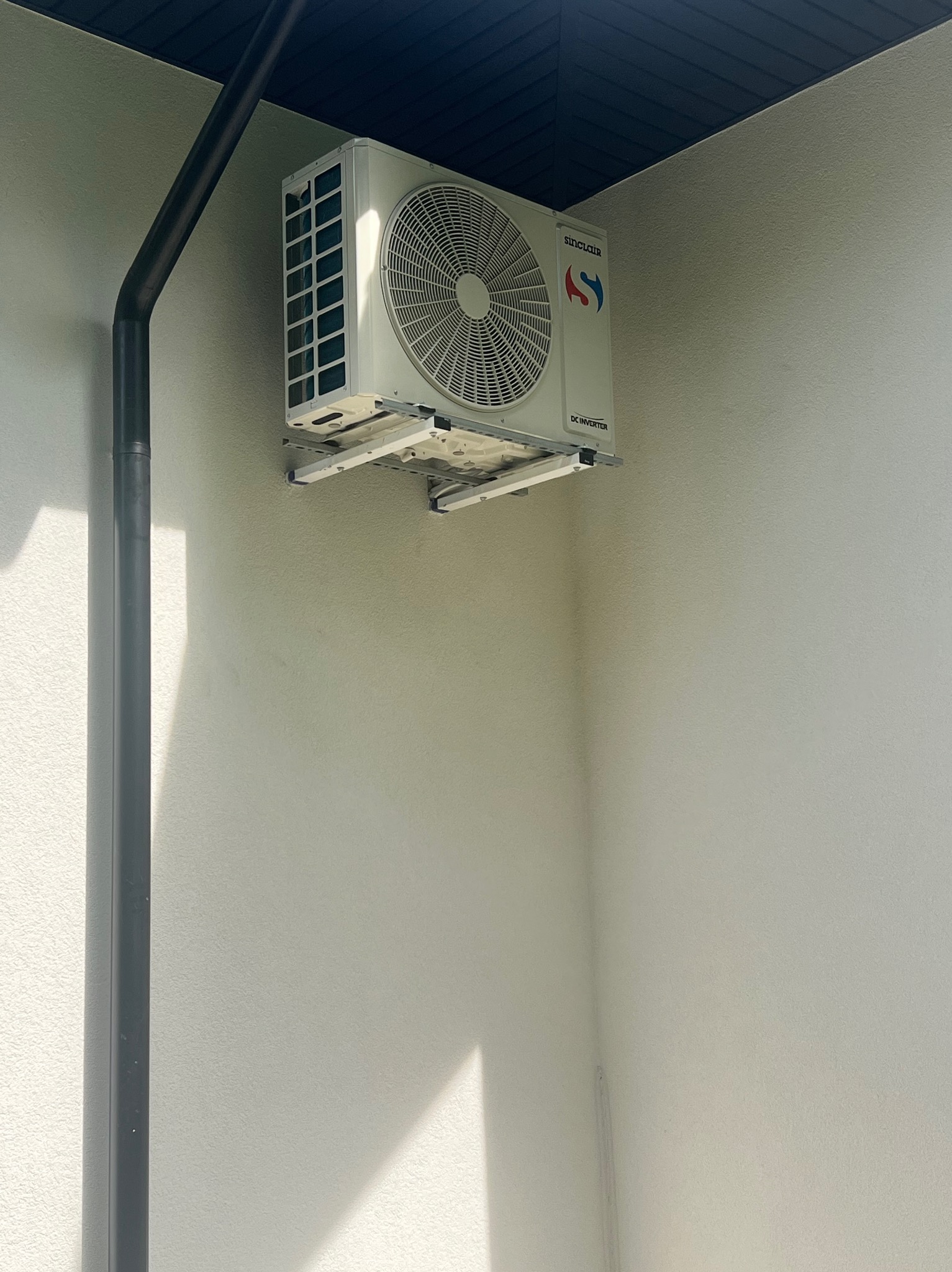 Zamontowana klimatyzacja Sinclair DC Inverter na białej ścianie budynku, z widoczną rurą spustową i fragmentem dachu. Widoczne mocowania klimatyzacji.