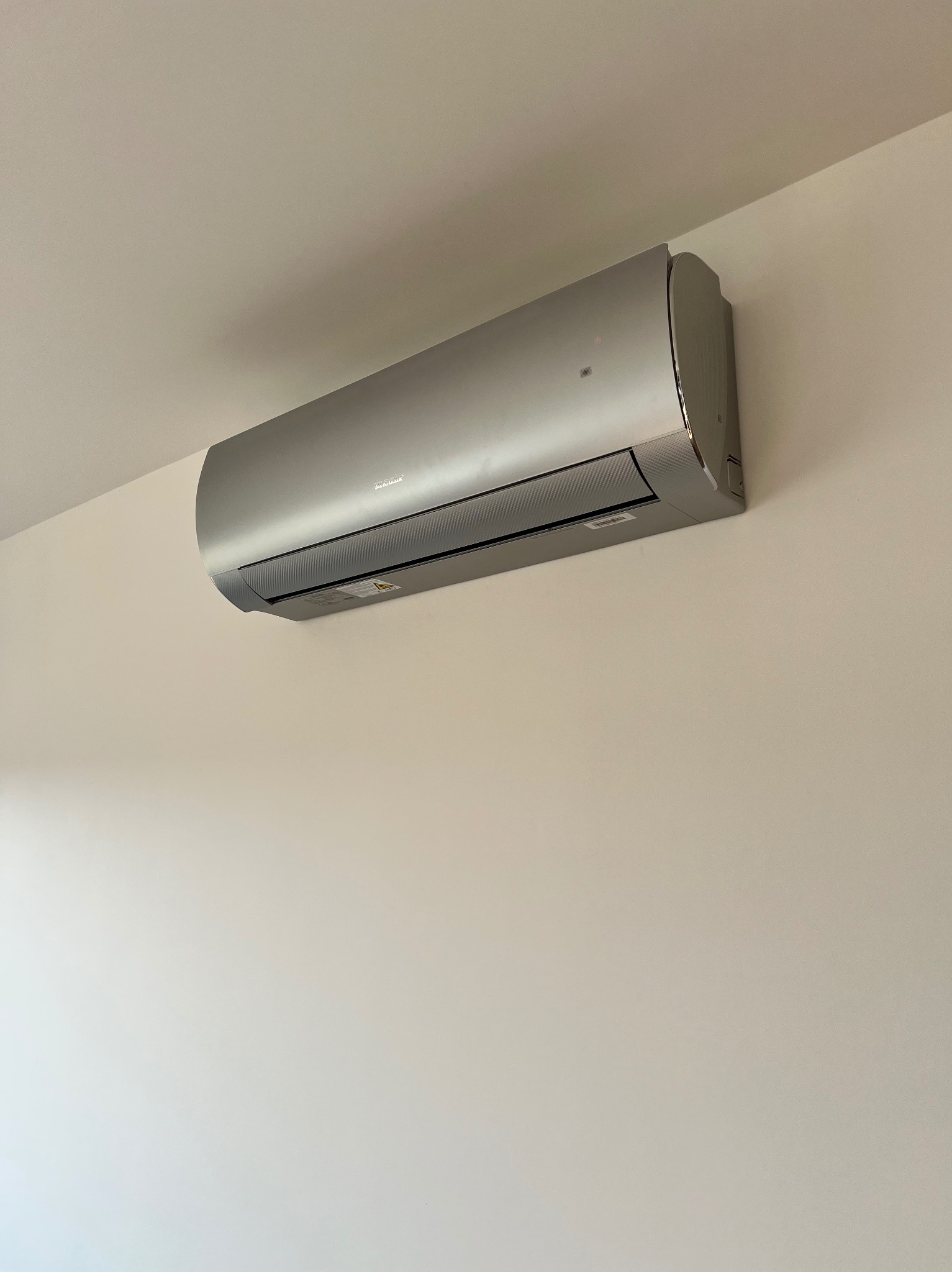 Zamontowany na ścianie, srebrny klimatyzator z poziomymi żaluzjami, w minimalistycznym wnętrzu. Widok z dołu ukazuje gładką powierzchnię i subtelne detale wykończenia.