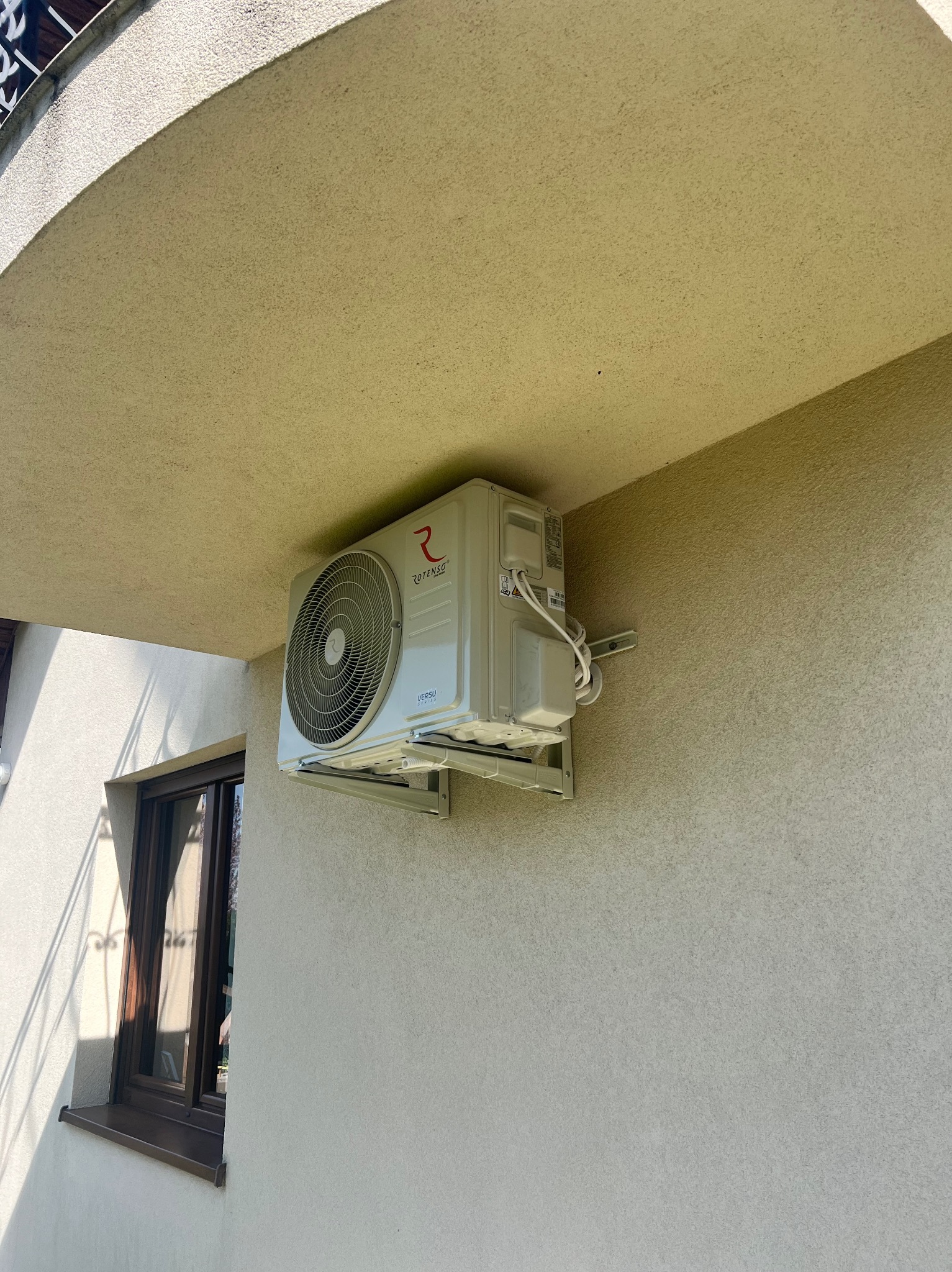 Zamontowana jednostka zewnętrzna klimatyzacji Rotenso na elewacji budynku z widocznym oknem. Biała obudowa, szara ściana, widoczne kable i wsporniki.