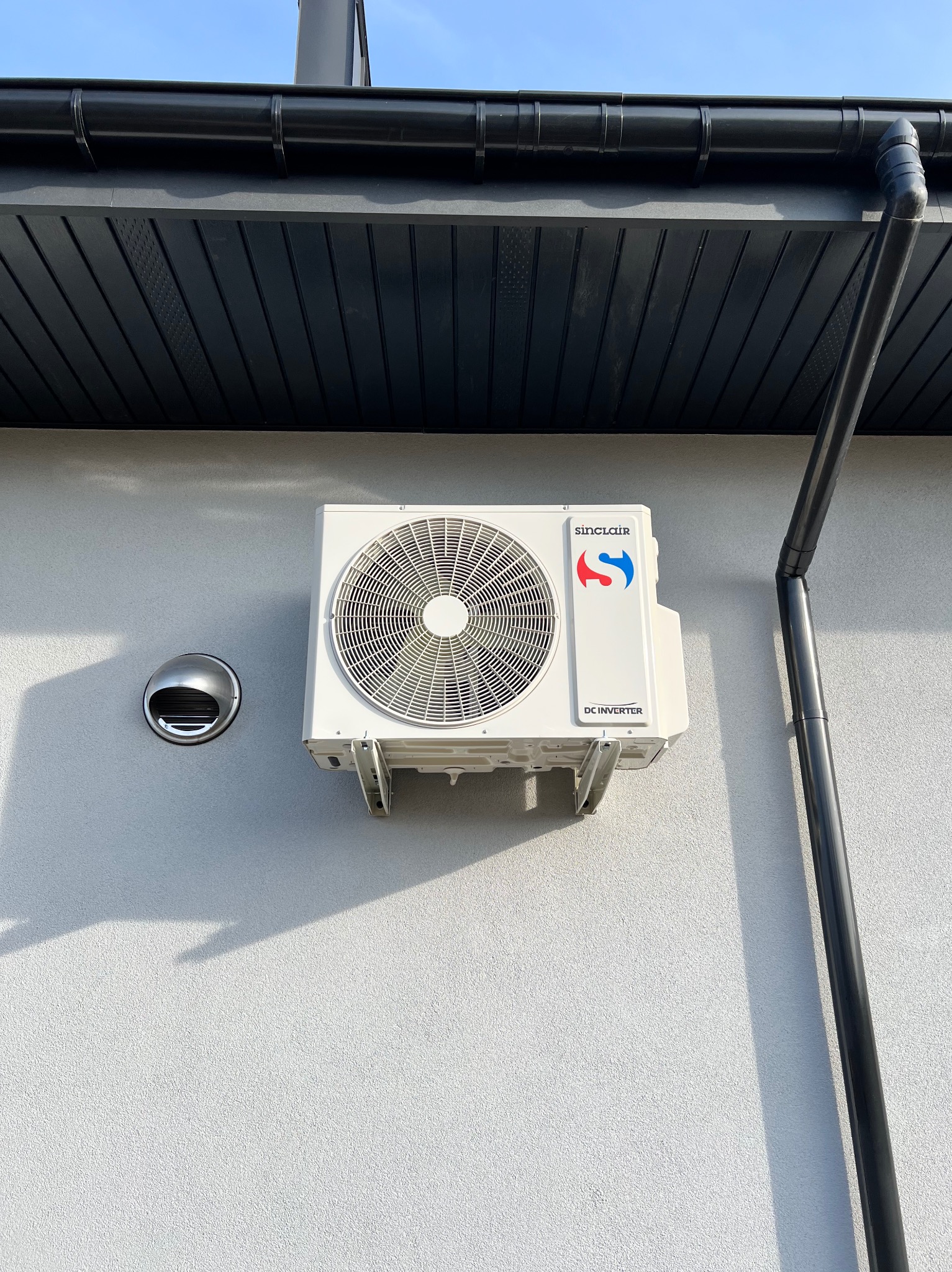 Biała jednostka zewnętrzna klimatyzacji Sinclair DC Inverter na szarej elewacji budynku, widok z dołu, obok rynna spustowa i kratka wentylacyjna.