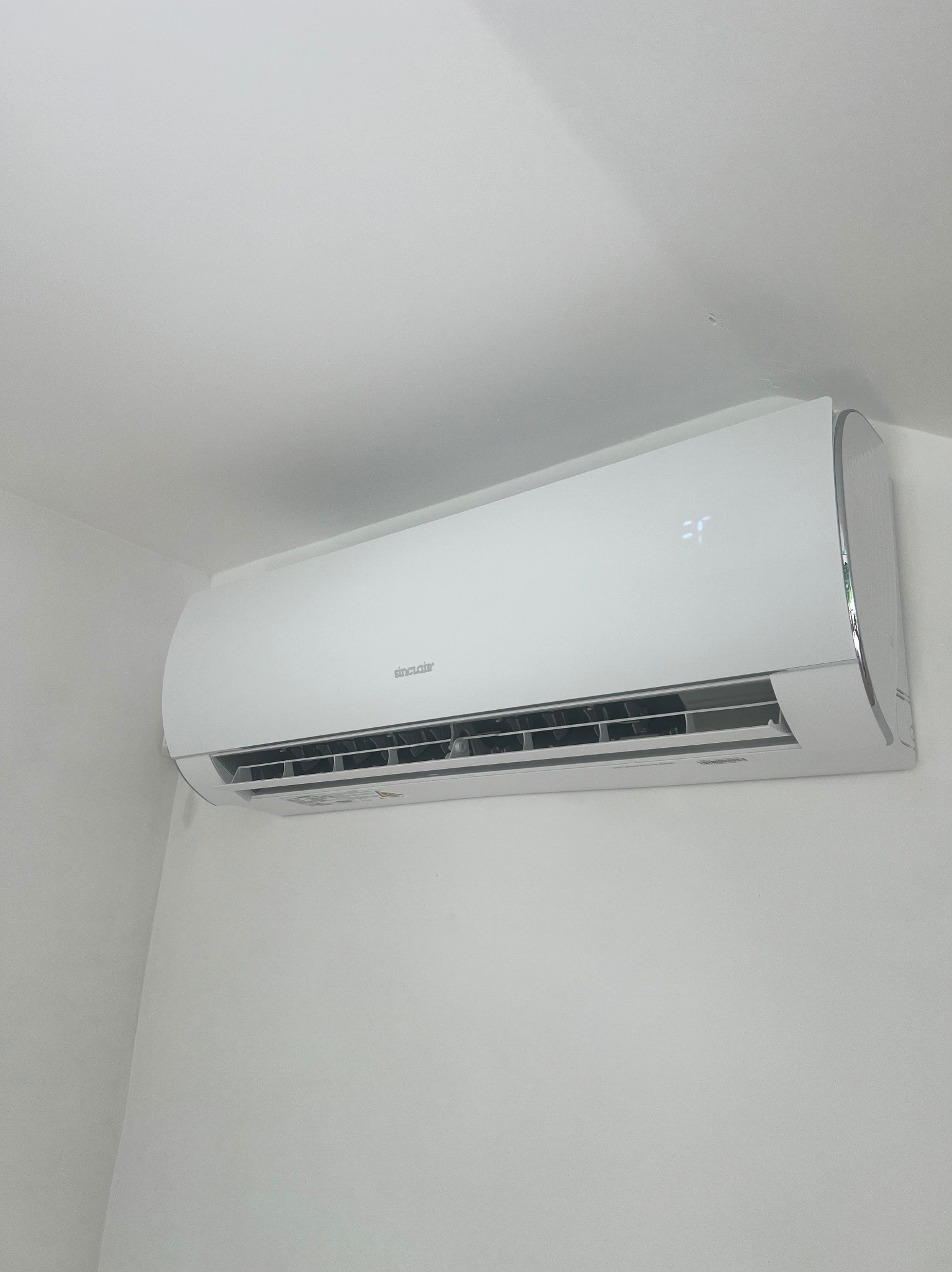 Biała klimatyzacja Sinclair, zamontowana na białej ścianie w rogu pomieszczenia. Widoczny wyświetlacz temperatury, żaluzje wlotu powietrza i obudowa urządzenia.