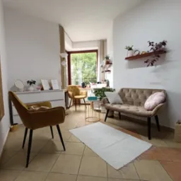 Jasne wnętrze gabinetu z sofą, krzesłami i roślinami. Na ścianie tablica z ilustracjami emocji. Przytulna aranżacja sprzyja rozmowie i relaksacji.