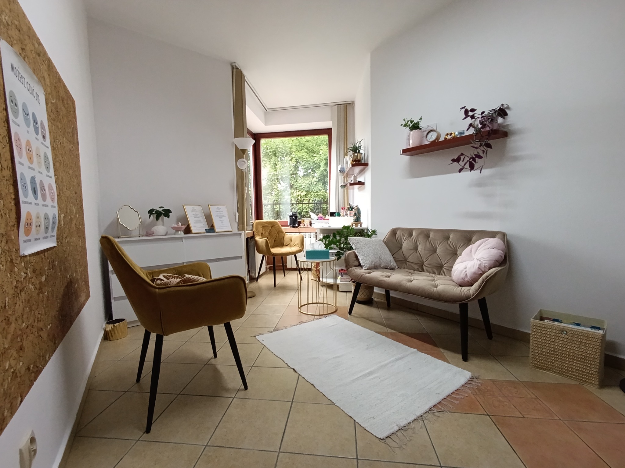 Jasne wnętrze gabinetu z sofą, krzesłami i roślinami. Na ścianie tablica z ilustracjami emocji. Przytulna aranżacja sprzyja rozmowie i relaksacji.