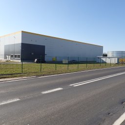 Biuro Obsługi Nieruchomości Dębscy Tadeusz Dębski - Duży budynek magazynowy z żółtym pasem w Żurominie, widziany z perspektywy ulicy. Obok zbiornik na wodę i ogrodzenie. Błękitne niebo w tle.