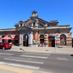 Biuro Obsługi Nieruchomości Dębscy Tadeusz Dębski - Elewacja budynku targowego z czerwonej cegły i szarego kamienia w słoneczny dzień. Widoczne stragany, auta i przechodnie. Charakterystyczna architektura.