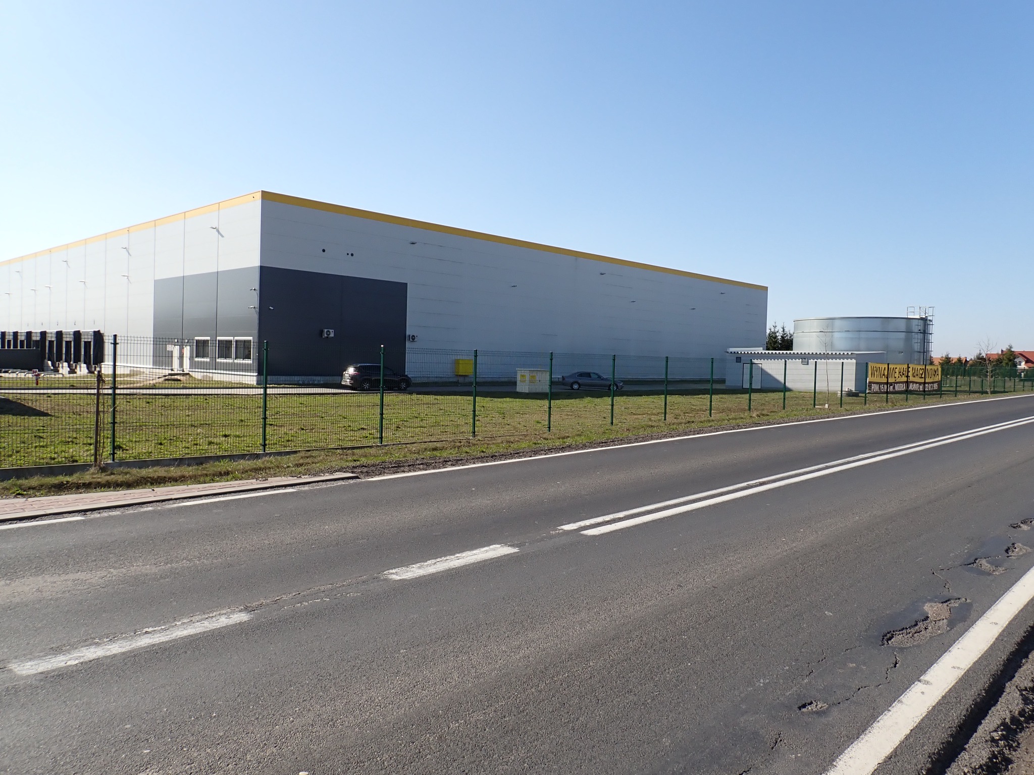 Duży budynek magazynowy z żółtym pasem w Żurominie, widziany z perspektywy ulicy. Obok zbiornik na wodę i ogrodzenie. Błękitne niebo w tle.