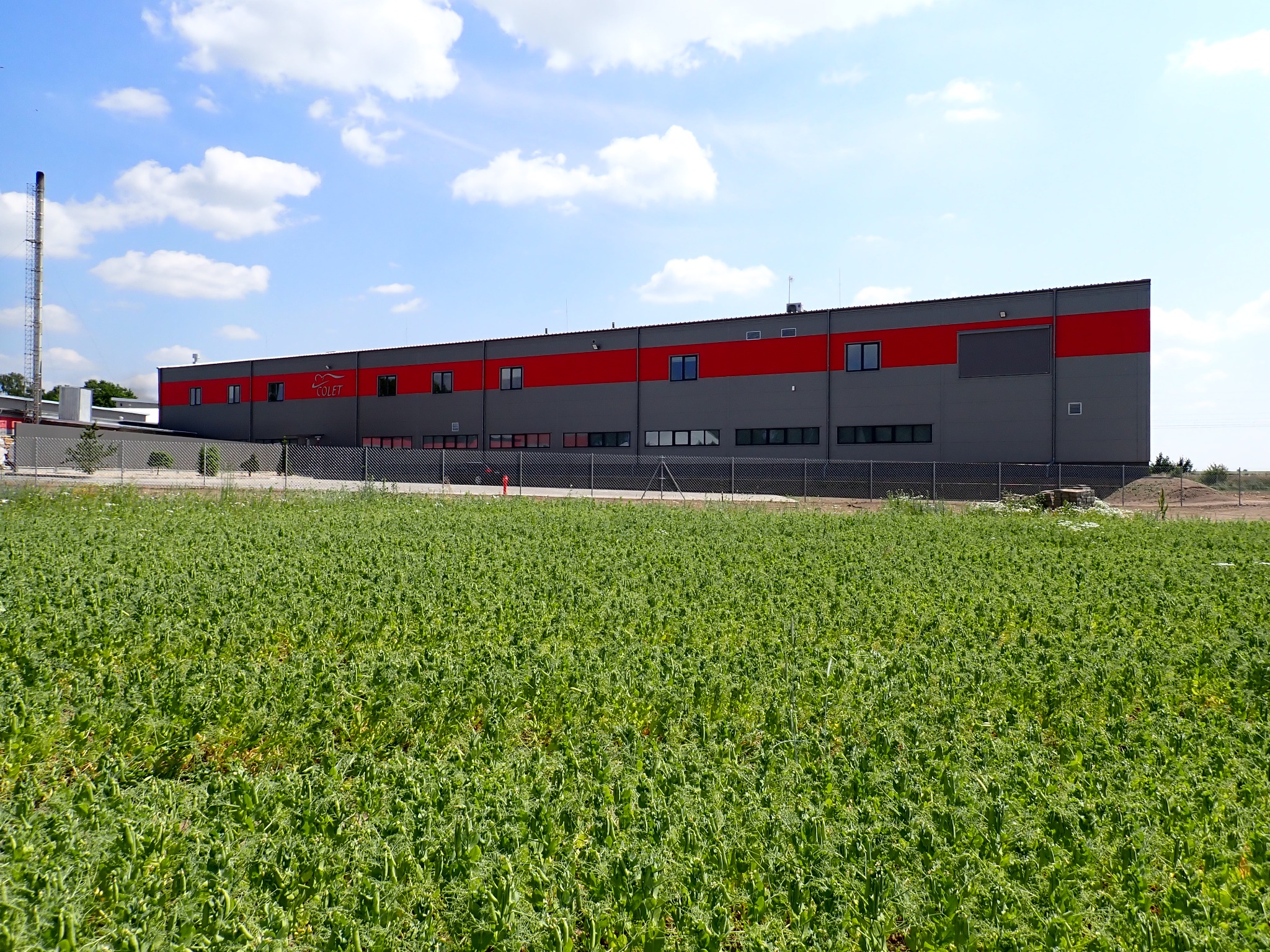 Szaro-czerwony budynek przemysłowy firmy Colet z kominem w tle, widziany z perspektywy pola uprawnego w słoneczny dzień z lekkim zachmurzeniem.