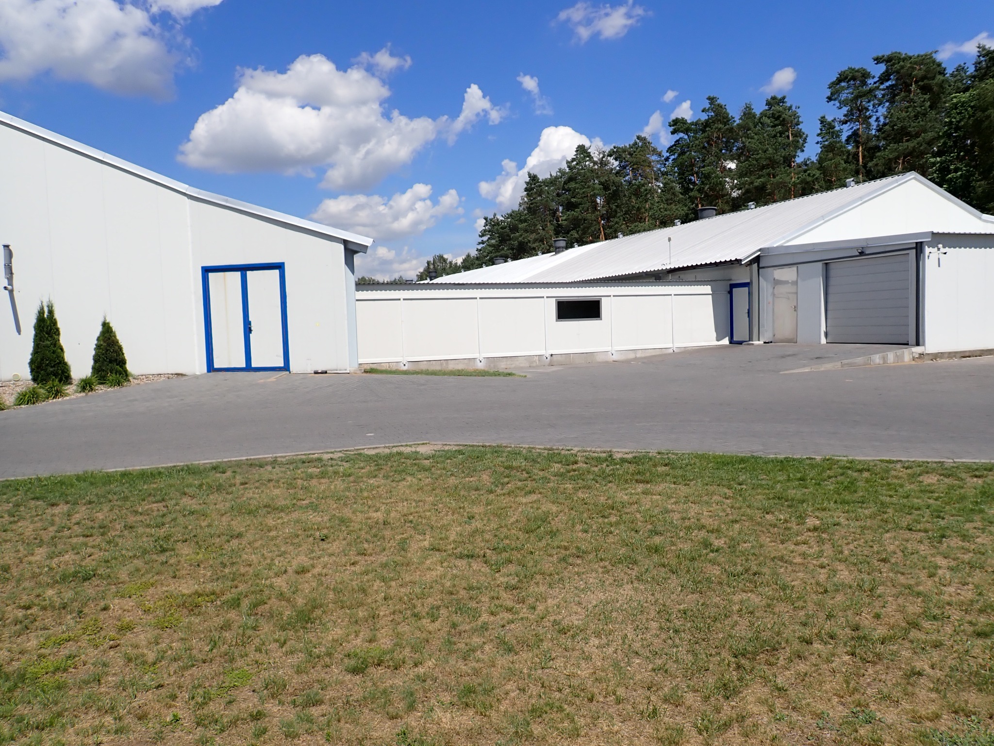 Białe budynki magazynowe z niebieskimi akcentami na tle błękitnego nieba z chmurami. Widok z zewnątrz na teren z trawą i drzewami w tle.