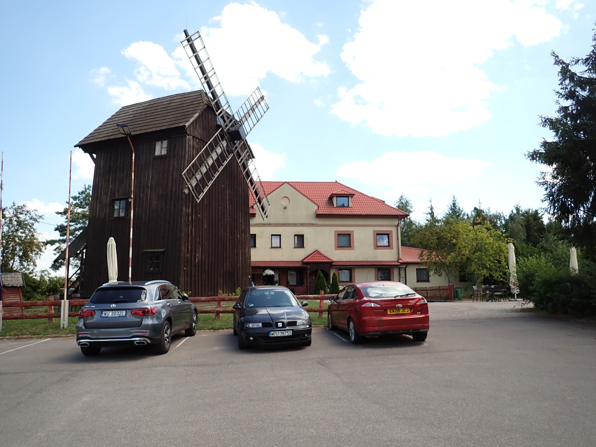 Malownicza sceneria z drewnianym wiatrakiem, budynkiem z czerwonym dachem i trzema samochodami na parkingu w słoneczny dzień.