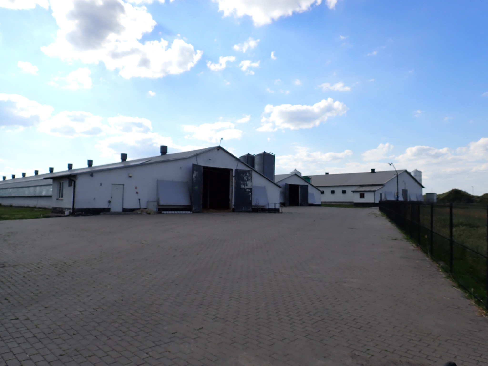 Rozległy kompleks budynków gospodarczych z otwartymi wrotami, widziany z brukowanego placu w słoneczny dzień. Widoczne silosy i ogrodzenie.
