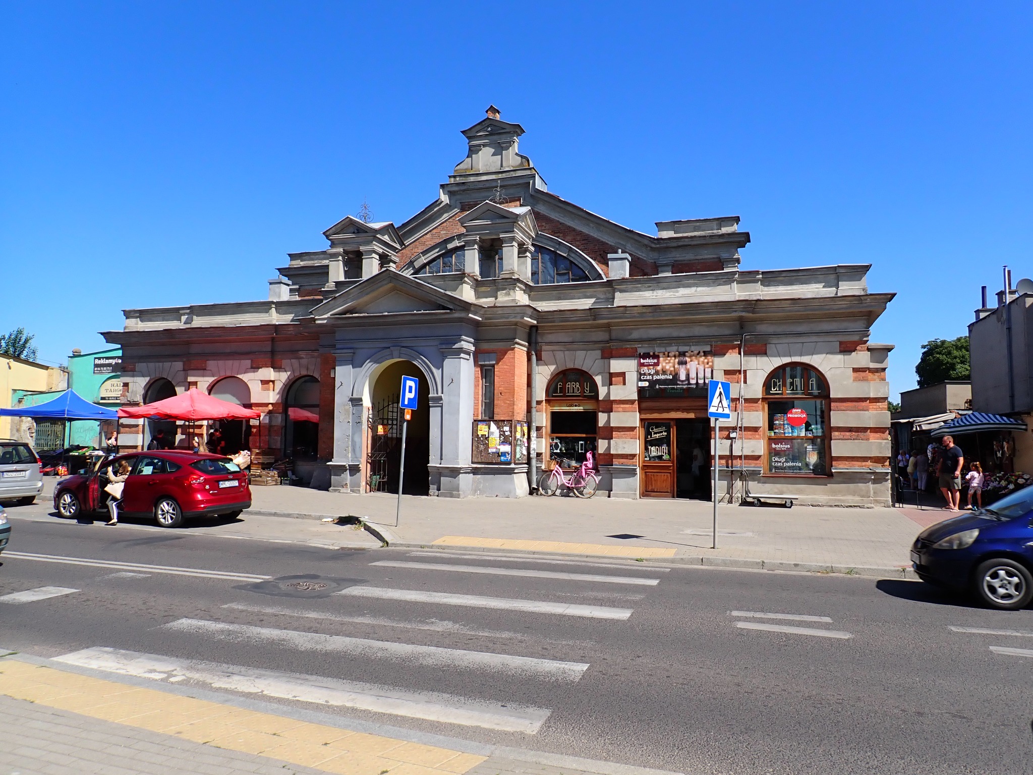 Elewacja budynku targowego z czerwonej cegły i szarego kamienia w słoneczny dzień. Widoczne stragany, auta i przechodnie. Charakterystyczna architektura.