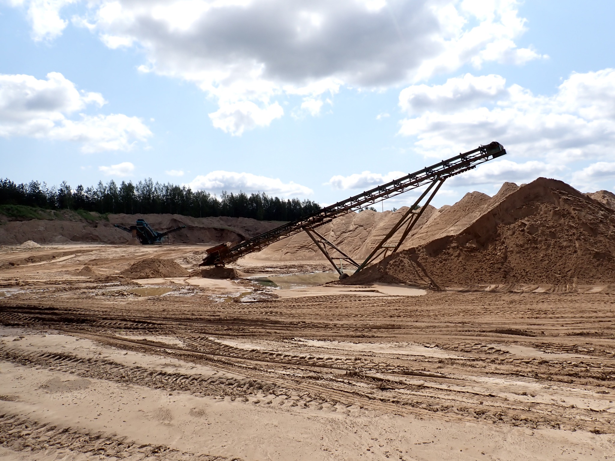Krajobraz kopalni piasku z taśmociągiem i śladami opon. W tle drzewa i błękitne niebo z chmurami. Ujęcie w słoneczny dzień.