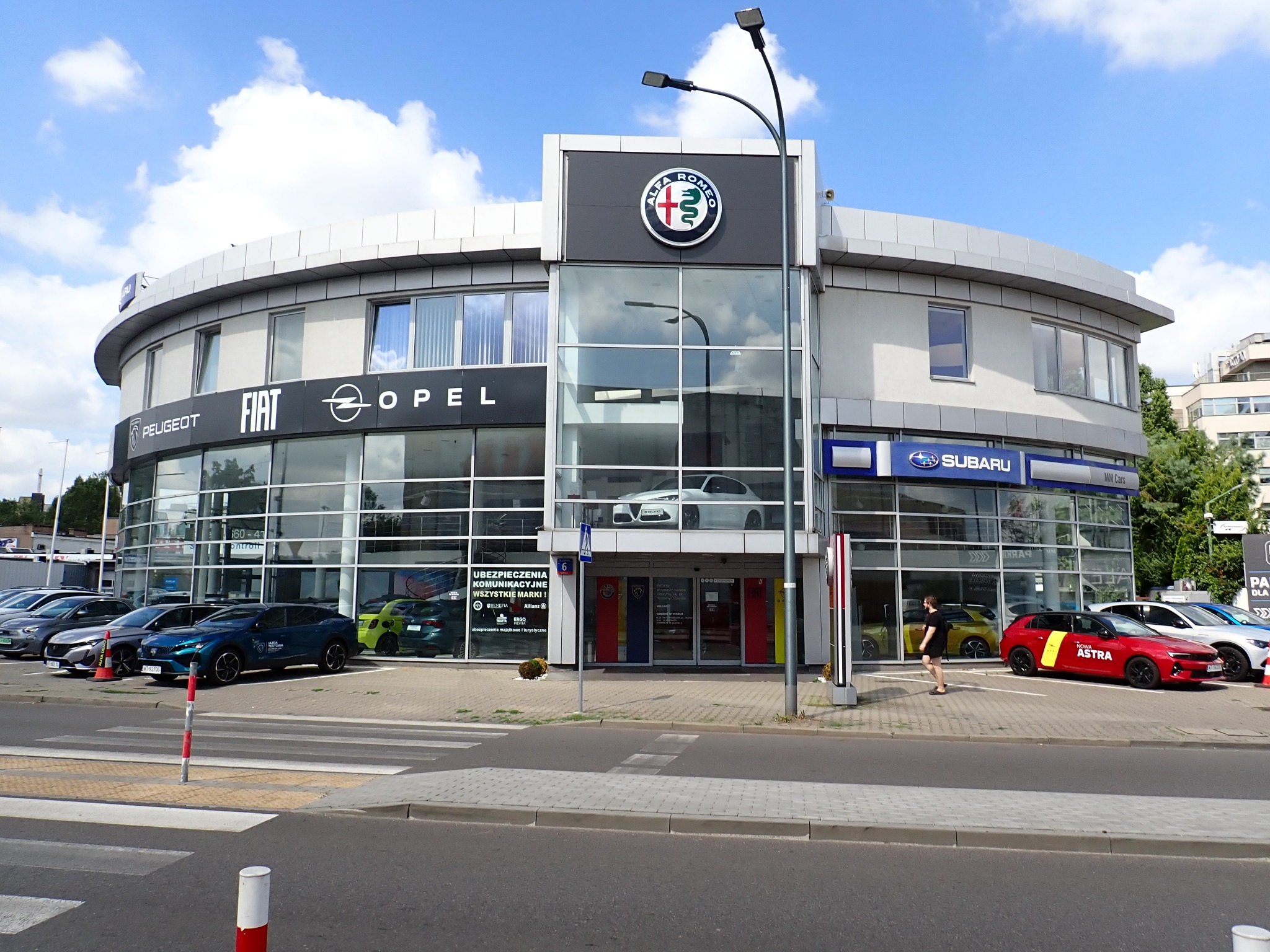 Salon samochodowy z logo Alfa Romeo, Fiat, Opel i Subaru. Na zewnątrz zaparkowane różne modele aut. Widoczny pieszy na chodniku. Słoneczny dzień.