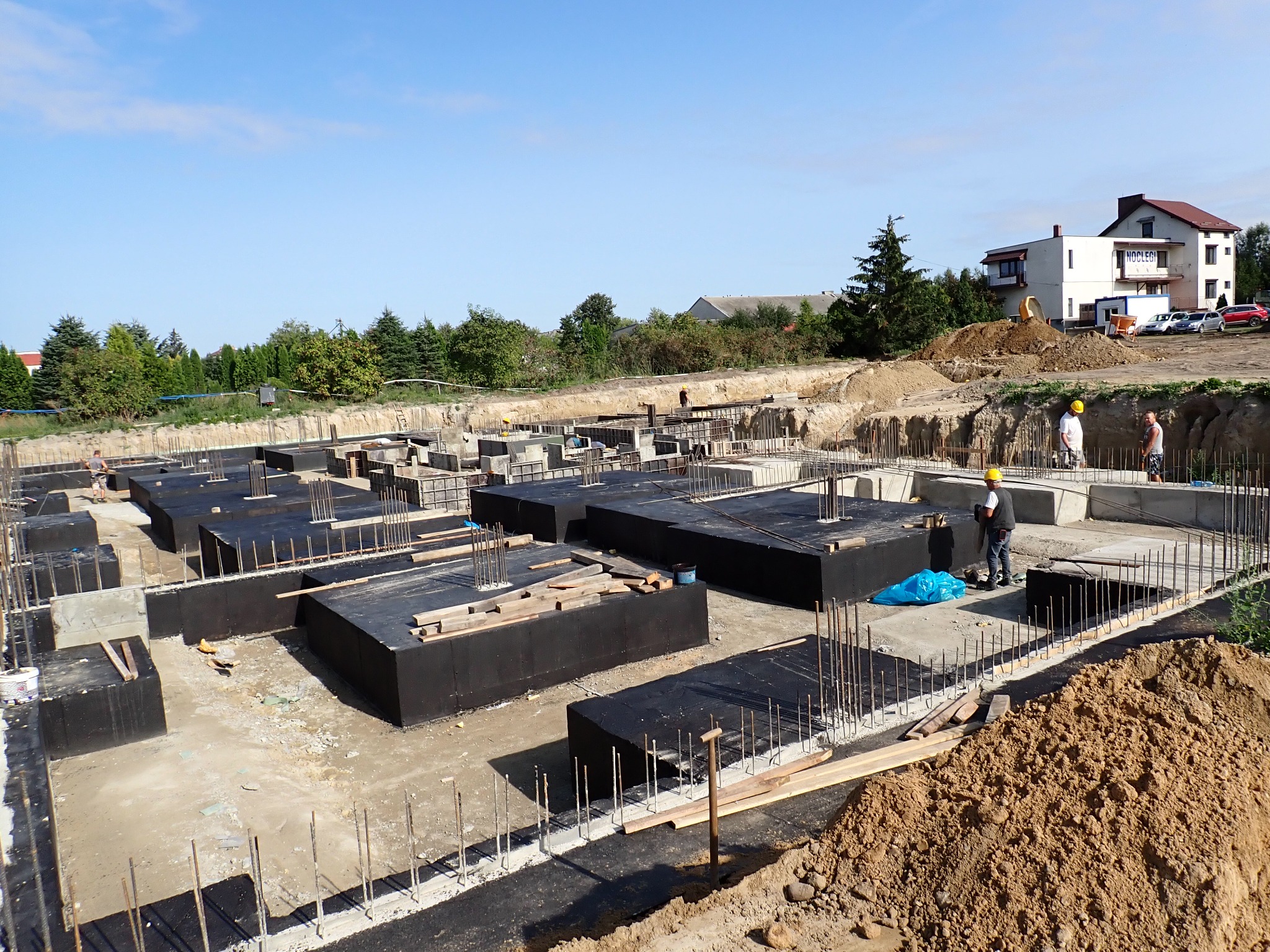 Fundamenty domu jednorodzinnego w trakcie budowy, zbrojenia i izolacja, widoczni pracownicy w kaskach, w tle budynek mieszkalny.