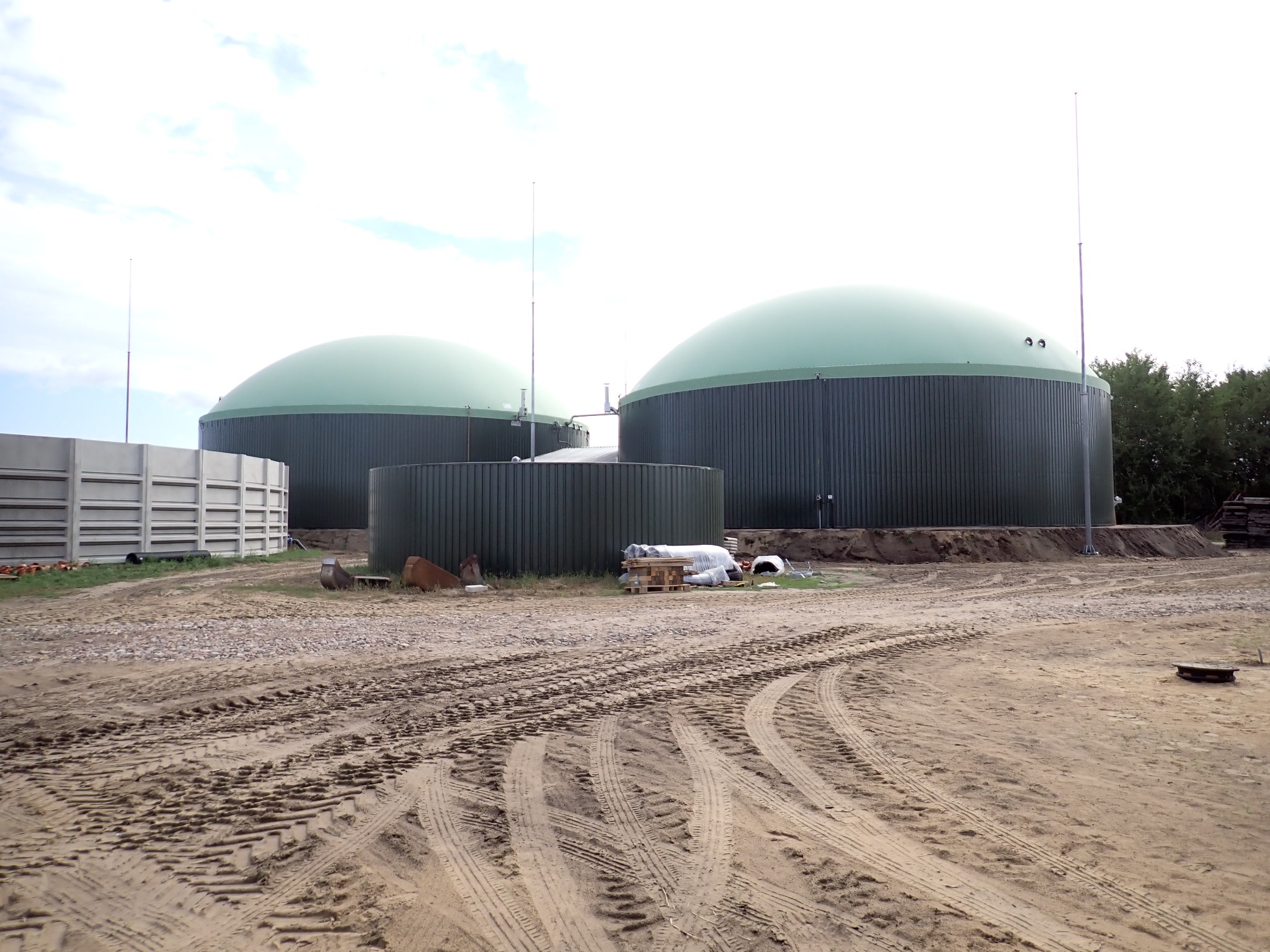 Dwa zbiorniki biogazu z zielonymi kopułami i antenami, otoczone ziemią z wyraźnymi śladami opon. Widoczne betonowe elementy i składowane materiały budowlane.