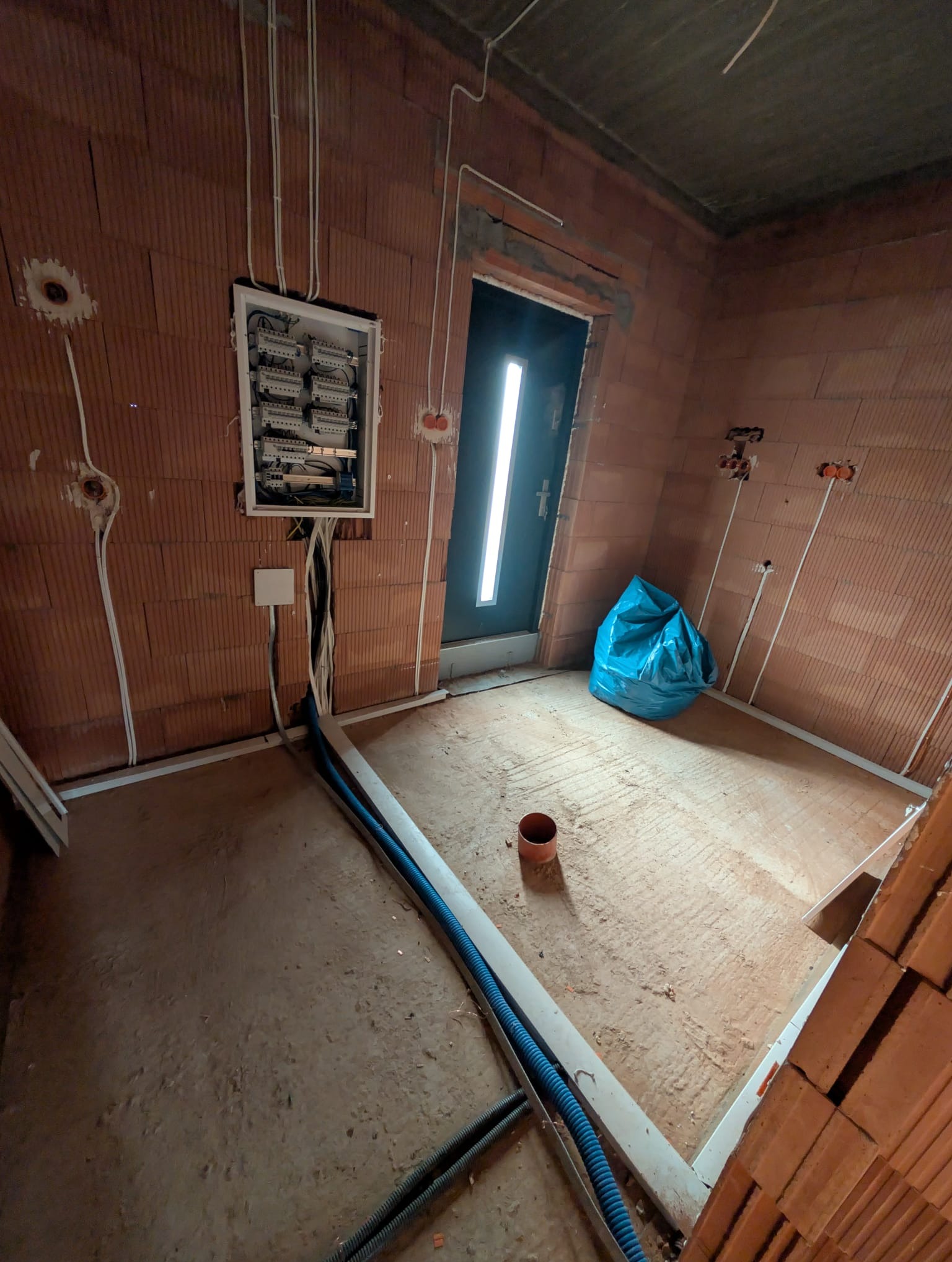 Wnętrze w trakcie instalacji elektrycznej: rozdzielnia, kable w peszlach, puszki w ścianach z cegły dziurawki. Niebieski worek na gruz przy drzwiach.
