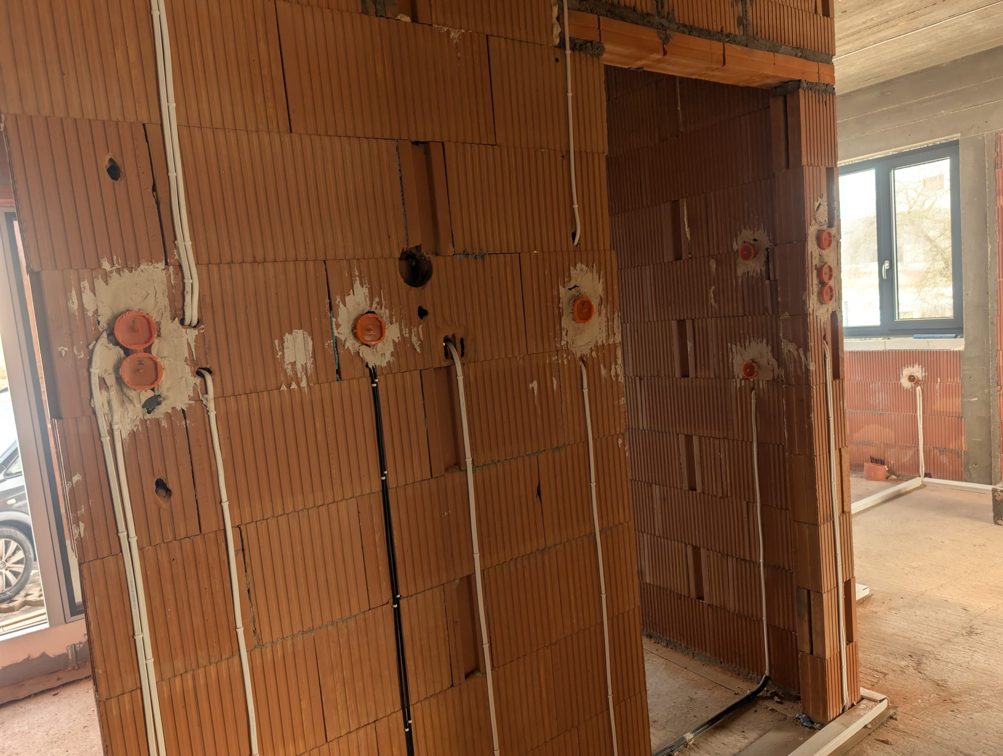 Wnętrze w trakcie budowy z widocznymi przewodami elektrycznymi w ścianach z cegły dziurawki i pomarańczowymi puszkami instalacyjnymi. Widoczne okno i surowy beton.