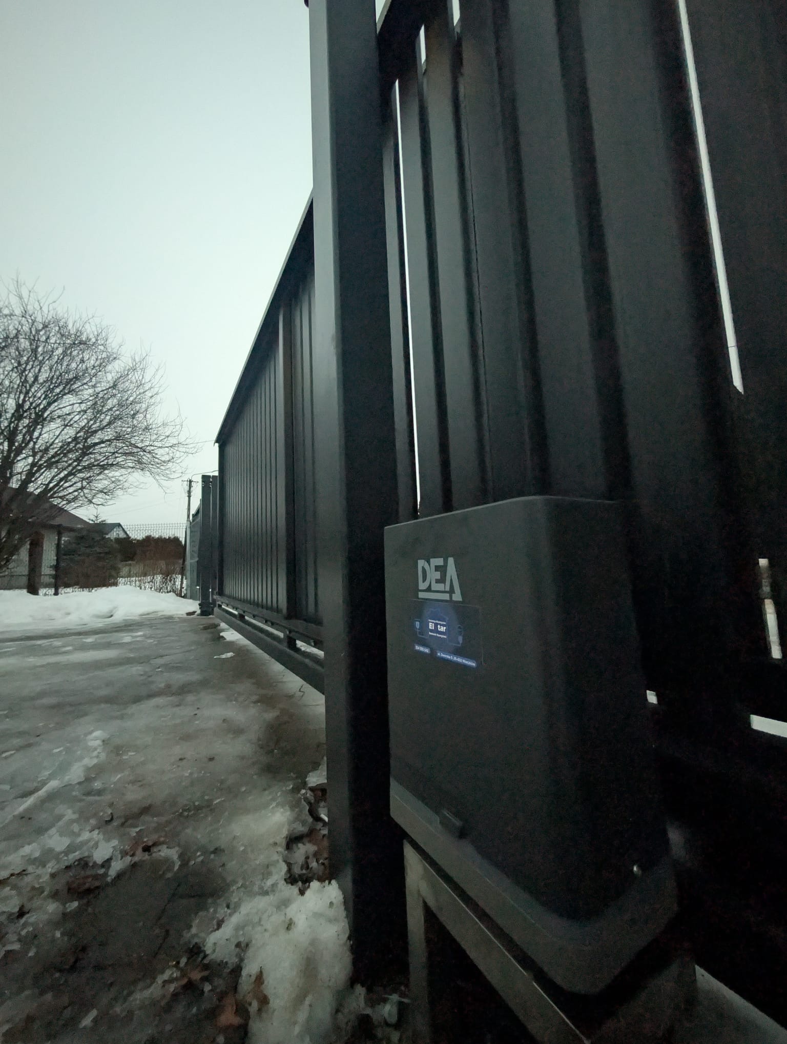 Automatyczna brama przesuwna z napędem DEA, widoczna w zimowej scenerii. Metalowa konstrukcja bramy, częściowo pokryta śniegiem, z widocznym mechanizmem sterującym.