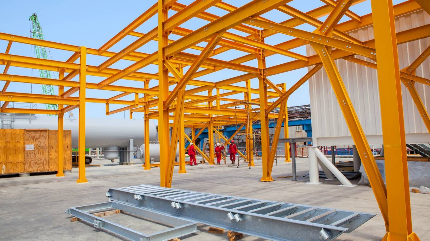 Żółta stalowa konstrukcja hali w trakcie budowy, z pracownikami w czerwonych kombinezonach w tle. Widoczny dźwig i elementy konstrukcyjne na pierwszym planie.