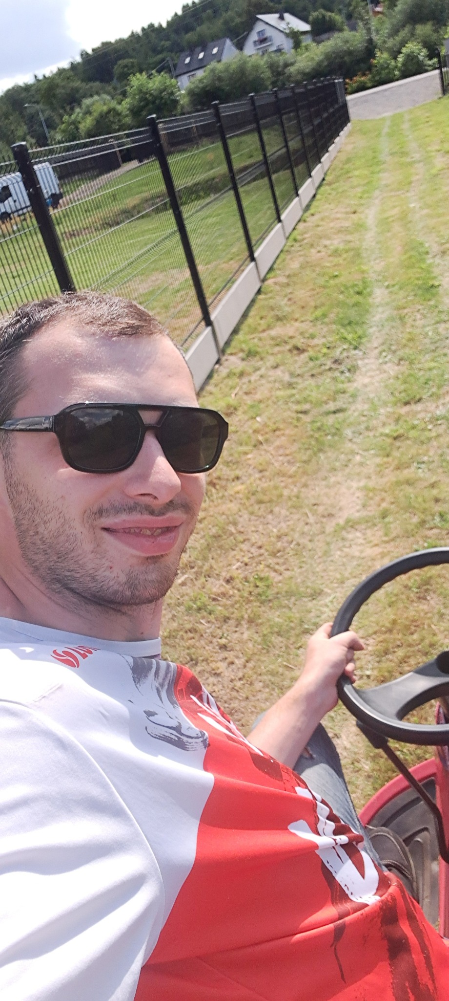 Uśmiechnięty mężczyzna w okularach słonecznych, siedzący na czerwonym traktorku do koszenia trawy, z widocznym ogrodzeniem i zielenią w tle.