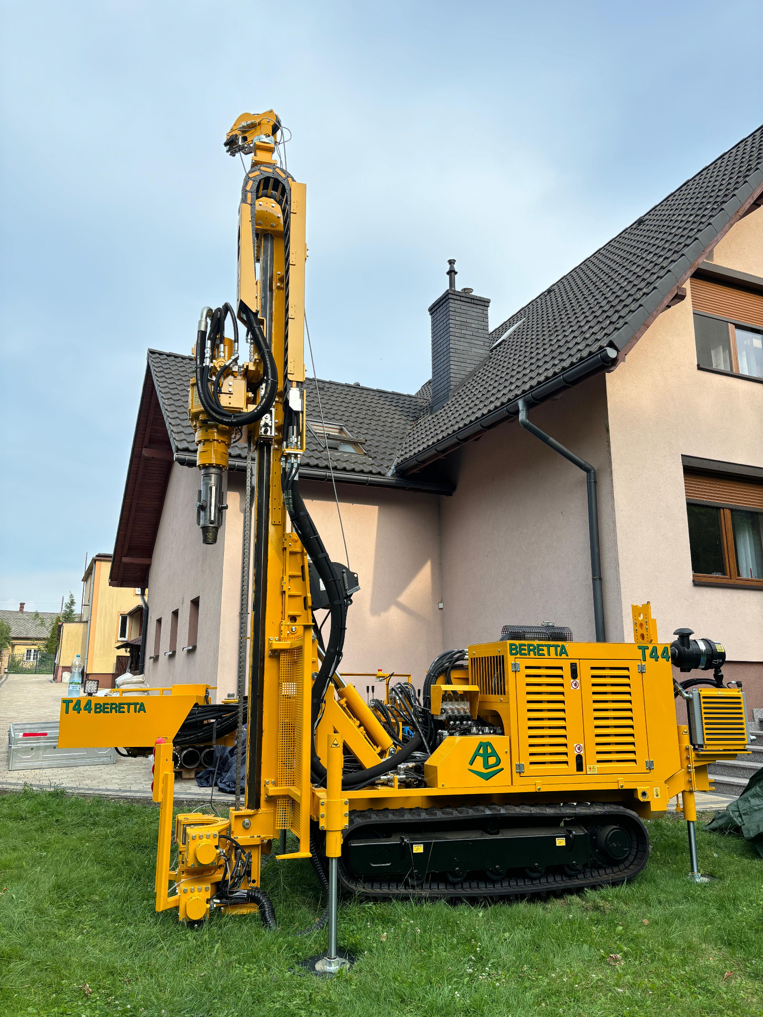 Żółta wiertnica Beretta T44 na trawie przed domem z ciemną dachówką i kominem. Maszyna gotowa do pracy, z wysuniętymi podporami stabilizującymi.