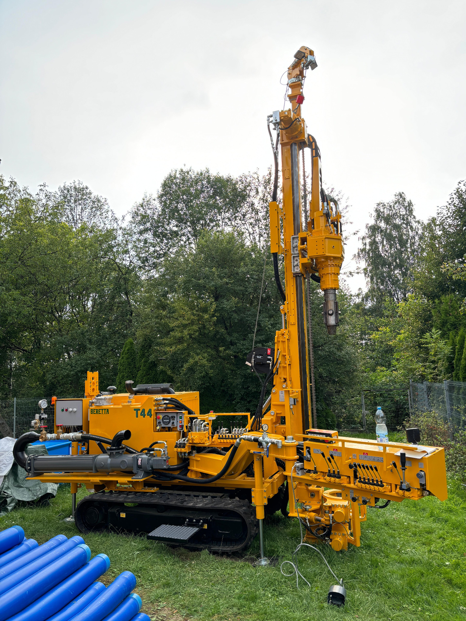 Żółta, gąsienicowa wiertnica Beretta T44 na trawie, z niebieskimi rurami w tle. Widok na maszynę z boku na tle drzew i pochmurnego nieba.