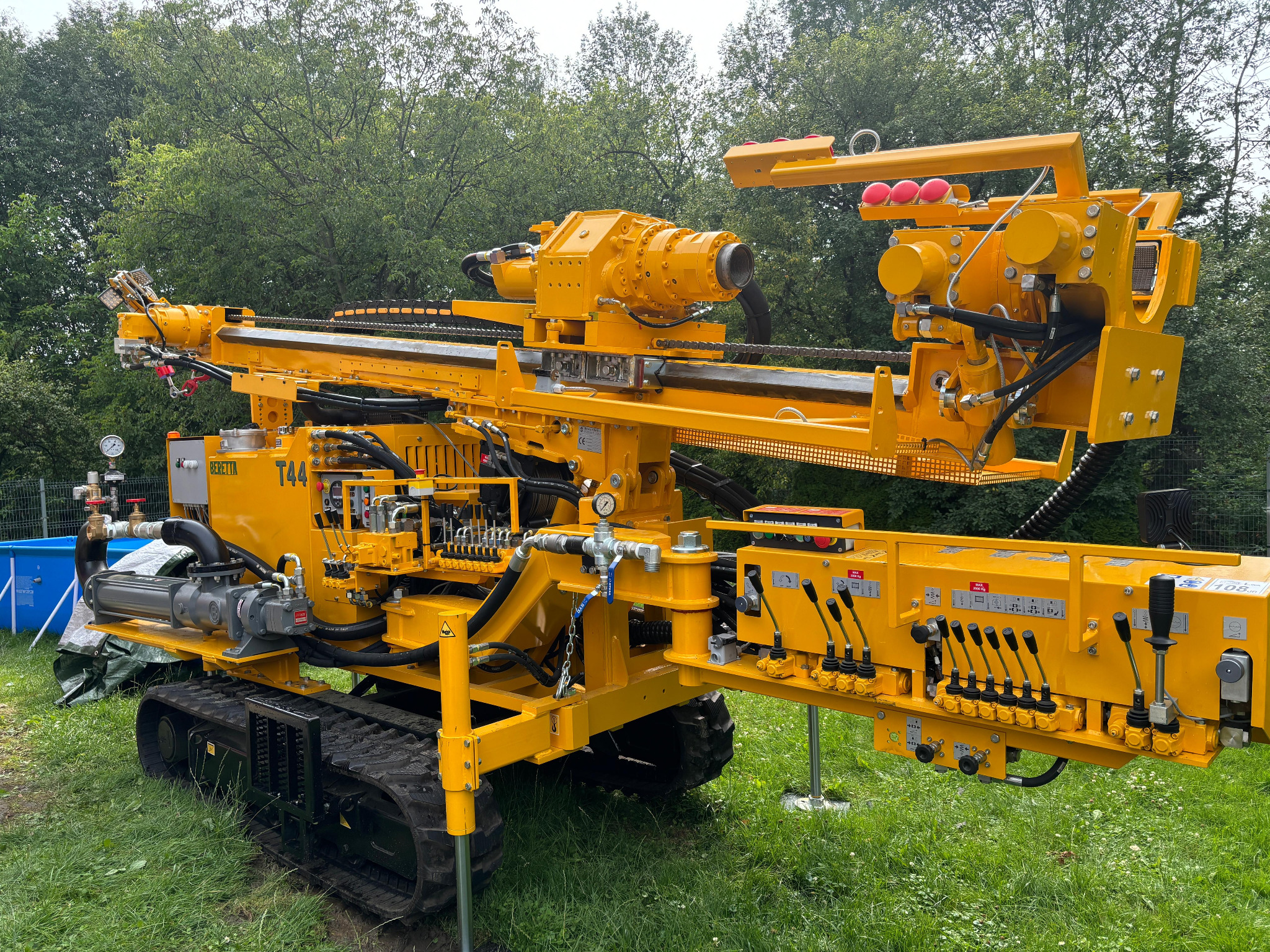 Żółta wiertnica Beretta T44 na gąsienicach, ustawiona na trawie, z widocznymi elementami hydraulicznymi i sterującymi. W tle zieleń drzew i fragment basenu.