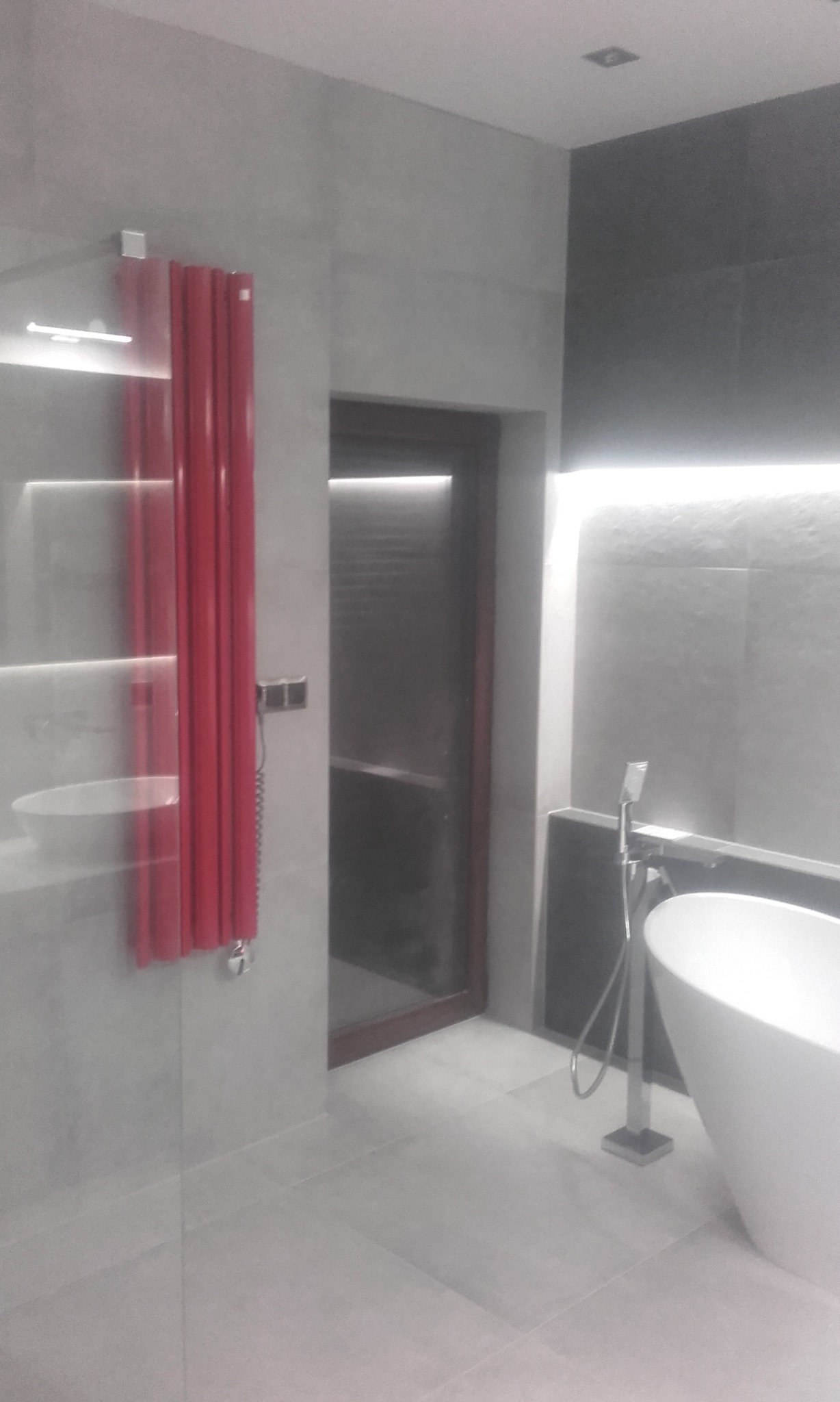 Nowoczesna łazienka z szarą płytką, czerwonym grzejnikiem drabinkowym i wolnostojącą białą wanną. Minimalistyczny design z oświetleniem LED.