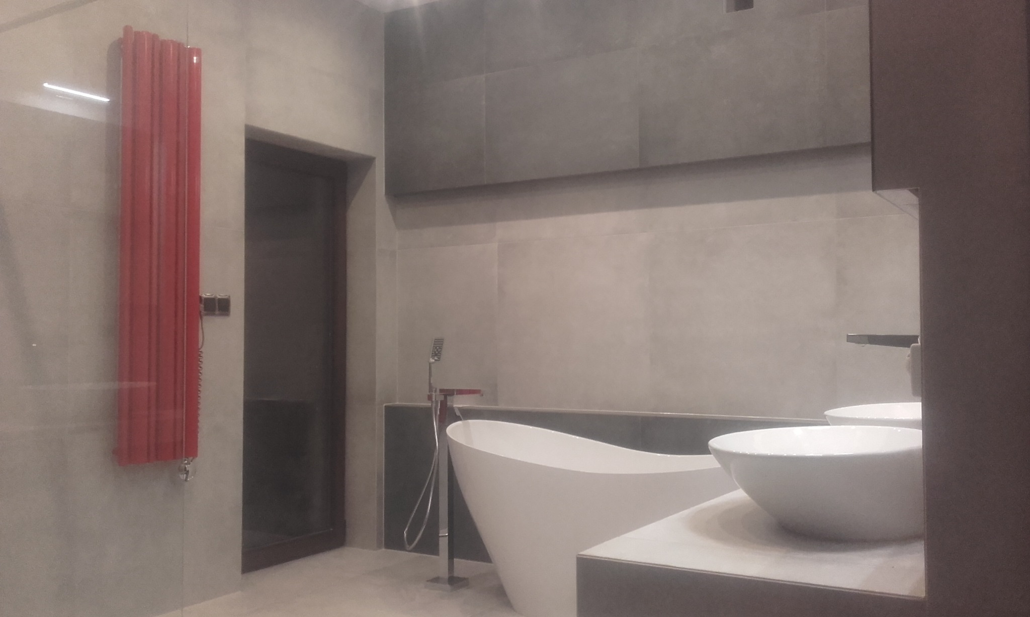 Nowoczesna łazienka z wolnostojącą wanną, umywalkami nablatowymi i czerwonym grzejnikiem dekoracyjnym na tle szarych płytek.