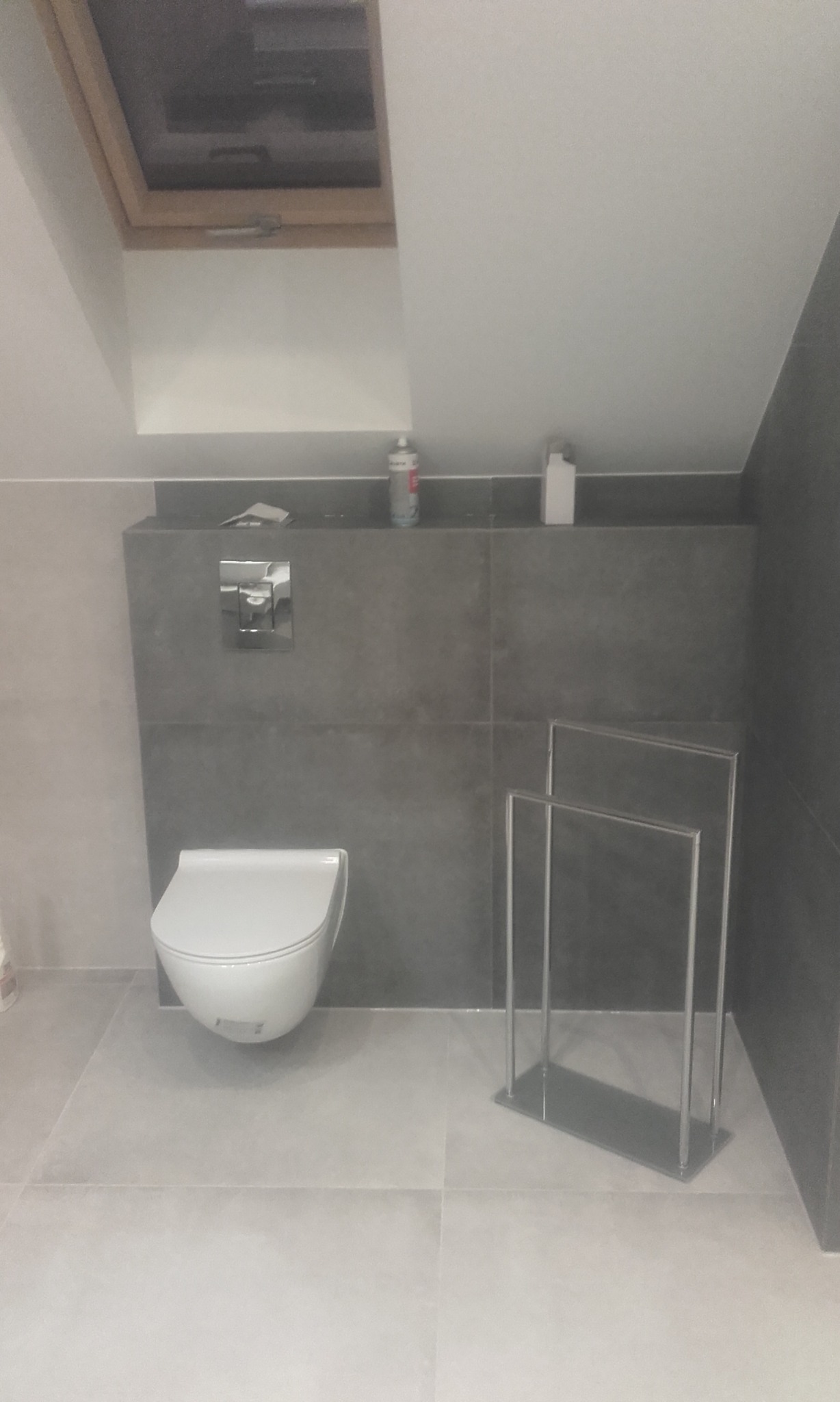 Nowoczesna łazienka na poddaszu z wiszącą toaletą, szarą glazurą i chromowanym wieszakiem na ręczniki. Uwagę przykuwa okno dachowe w drewnianej ramie.