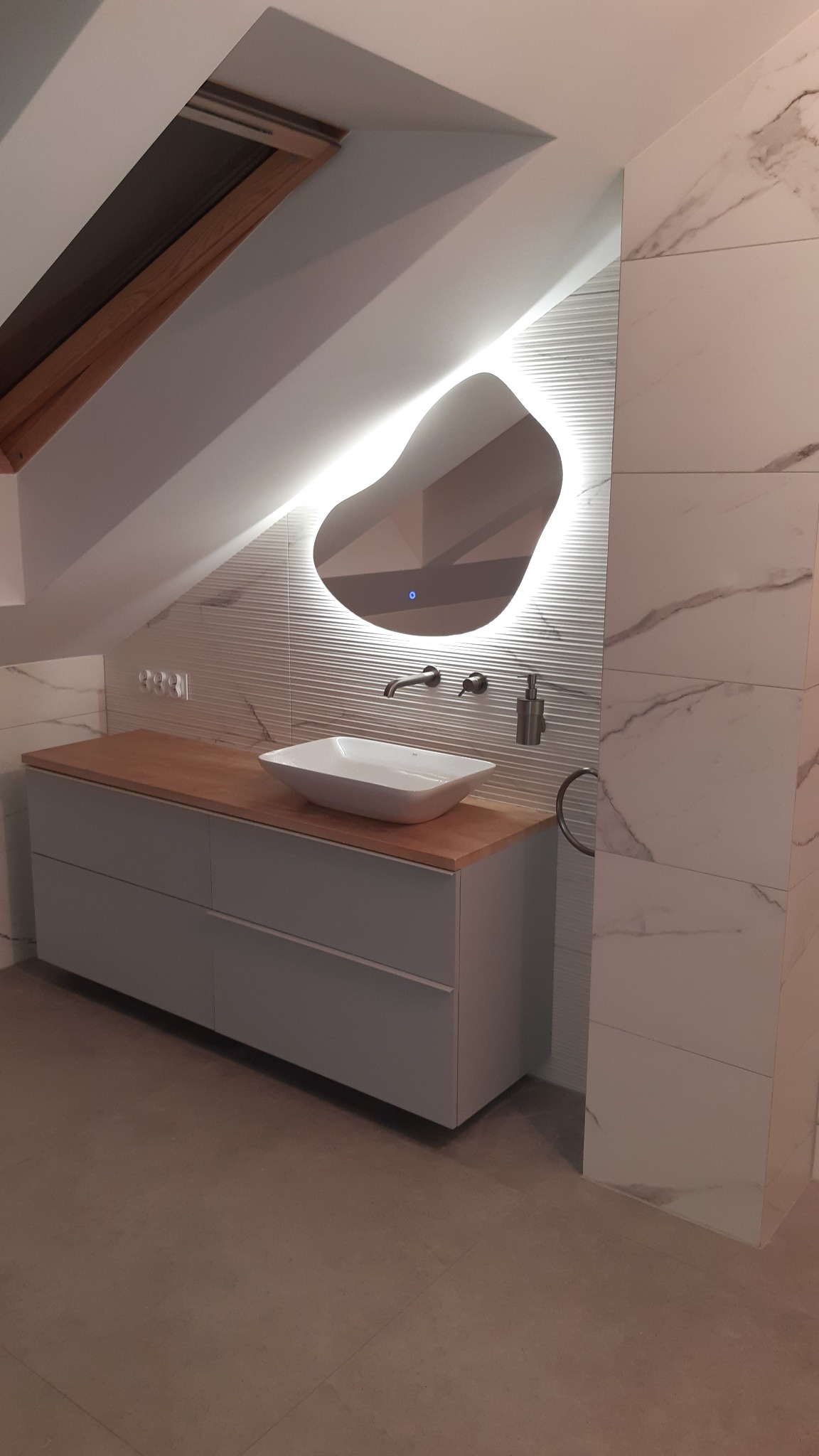 Nowoczesna łazienka na poddaszu z umywalką nablatową, podświetlanym lustrem o nieregularnym kształcie i marmurowymi płytkami. Ukośny sufit z oknem dachowym.