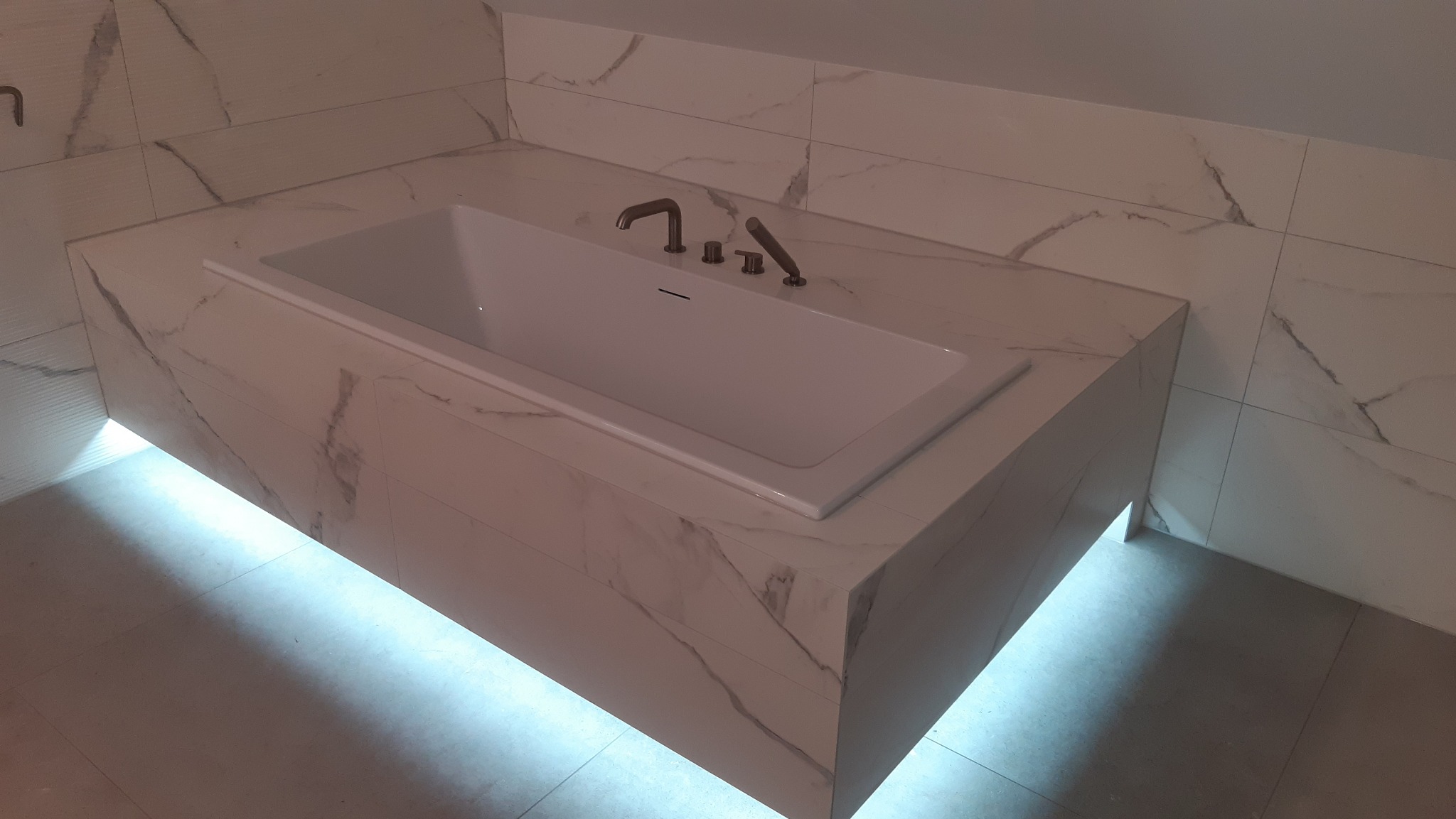 Nowoczesna wanna obudowana płytkami imitującymi marmur, z podświetleniem LED od dołu, tworząca efekt pływającej bryły w minimalistycznej łazience.