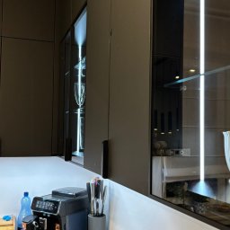 MEBLEMATE MATEUSZ RYNDAK - Nowoczesna kuchnia z czarnymi szafkami, oświetleniem LED wewnątrz, szklanymi frontami i blatem. Na blacie ekspres do kawy i akcesoria kuchenne.