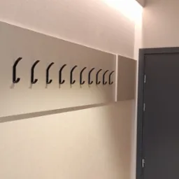 Nowoczesny wieszak ścienny z czarnymi haczykami na jasnej płycie, zamontowany w przedpokoju obok szarych drzwi z domofonem i oświetleniem LED na suficie.