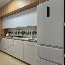 MEBLEMATE MATEUSZ RYNDAK - Nowoczesna kuchnia z białymi frontami i drewnianymi szafkami górnymi. Na blacie suszone zioła, obok biała lodówka Haier. Jasna podłoga z płytek.