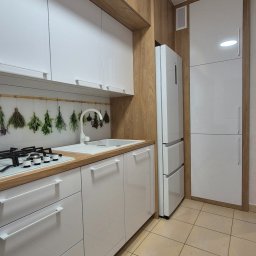 MEBLEMATE MATEUSZ RYNDAK - Nowoczesna kuchnia z białymi frontami i drewnianymi akcentami. Widoczny zlew, płyta gazowa i lodówka. Na ścianie dekoracja z suszonych ziół.