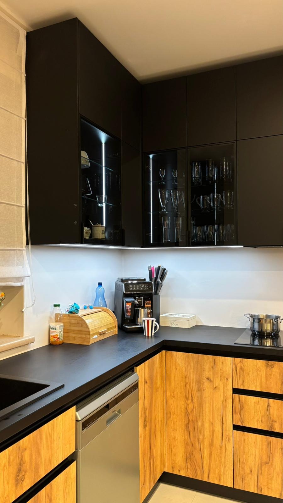 Nowoczesna kuchnia z czarnymi górnymi szafkami z podświetleniem LED i drewnopodobnymi dolnymi szafkami, czarny blat, ekspres do kawy i akcesoria.
