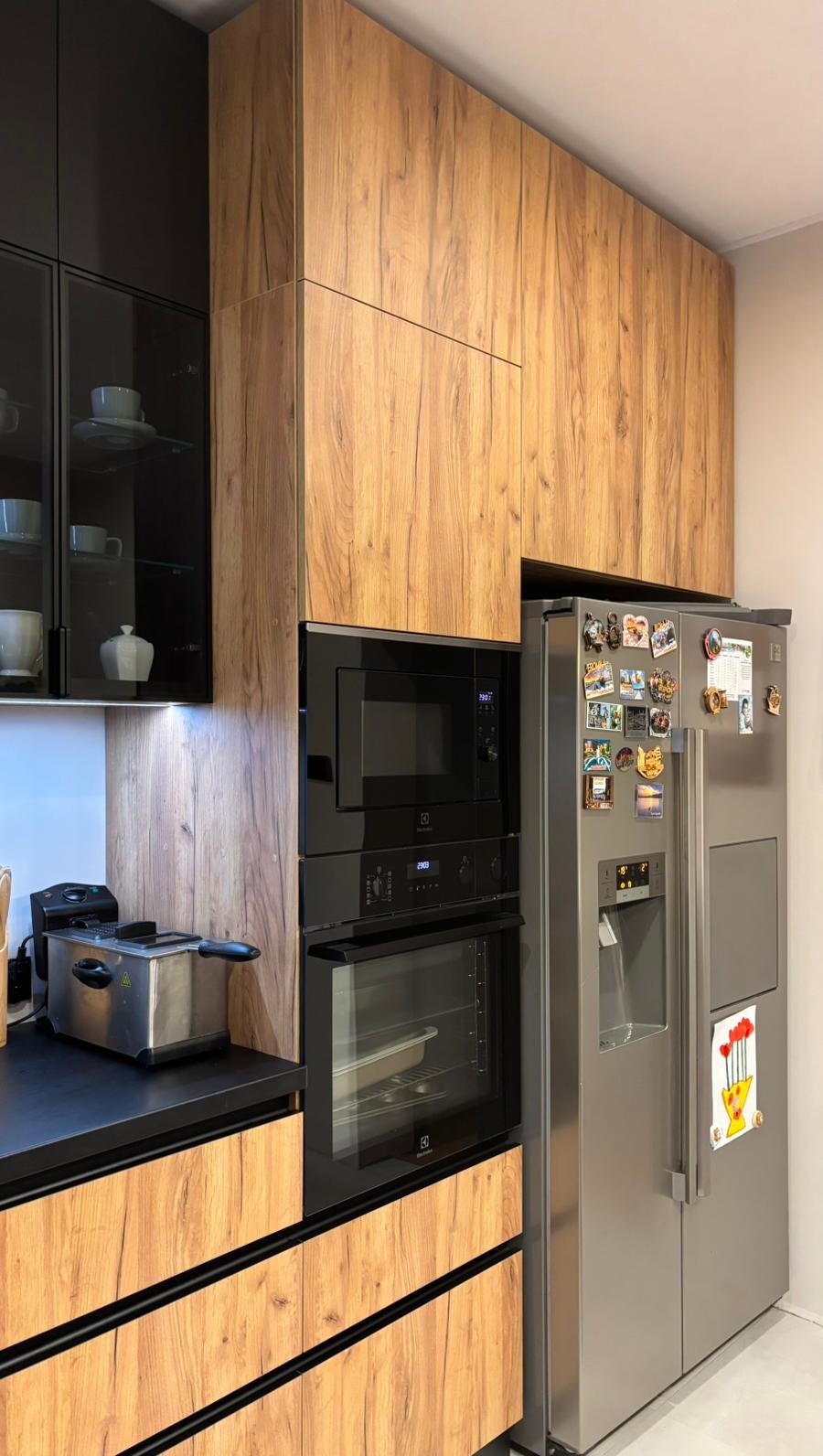 Nowoczesna kuchnia z drewnianymi frontami, czarnymi akcentami i sprzętem AGD: piekarnik, mikrofalówka i lodówka. Na blacie frytkownica.