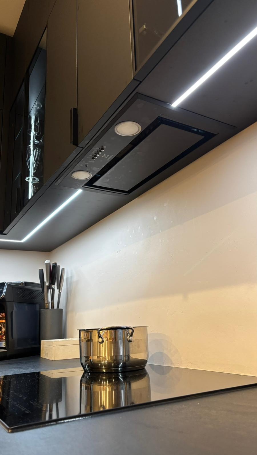 Czarny okap podszafkowy z oświetleniem LED, nad płytą indukcyjną z garnkami. Nowoczesna kuchnia z elementami szklanymi i sprzętem AGD.