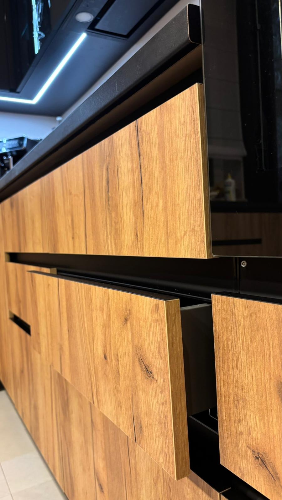 Szczegółowe ujęcie nowoczesnej kuchni z frontami w odcieniach drewna i czarnymi, minimalistycznymi uchwytami, z wysuniętą szufladą, ukazujące precyzję wykonania.