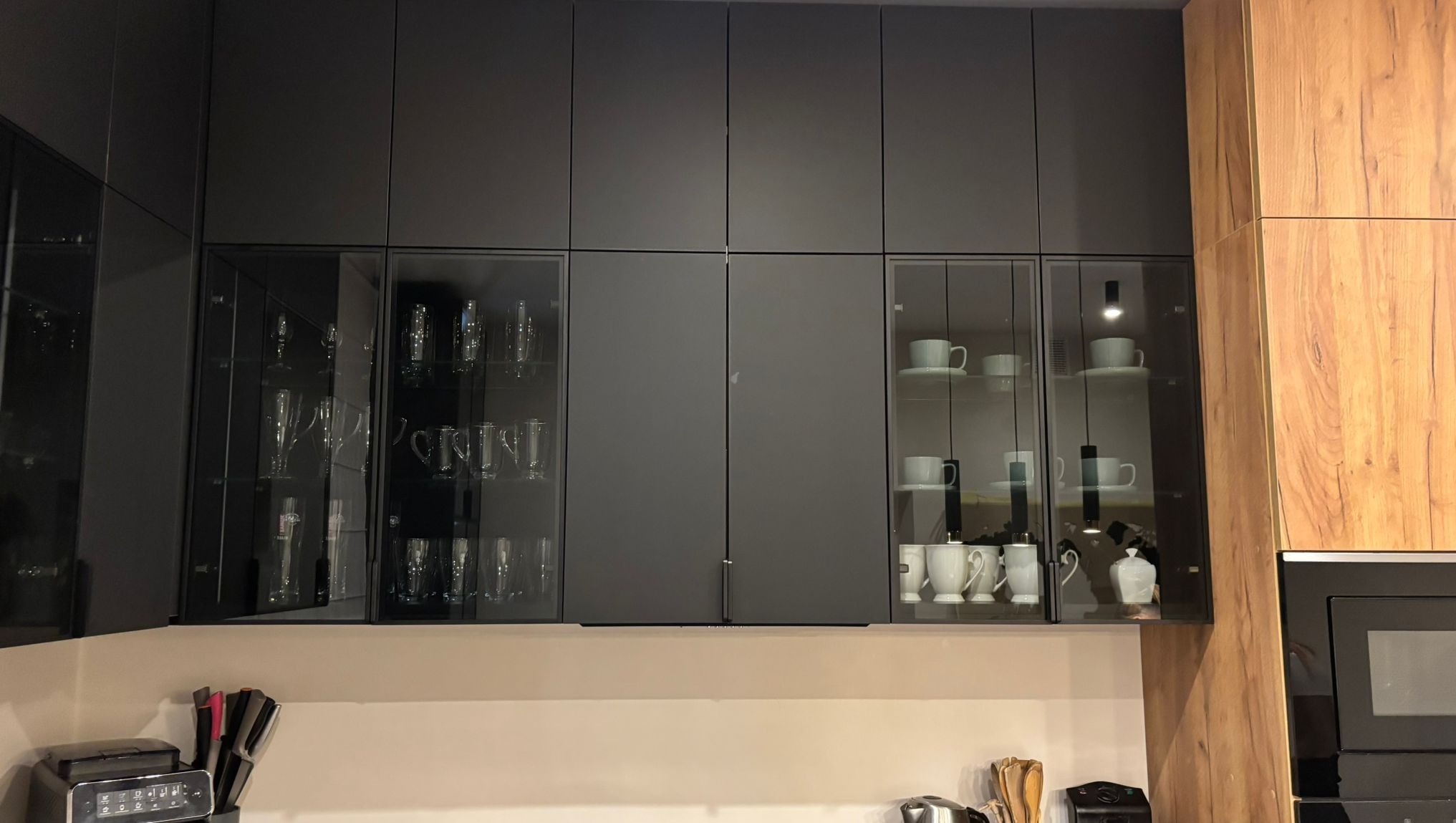 Nowoczesna zabudowa kuchenna z górnymi szafkami: szare, pełne fronty i przeszklone witryny z oświetleniem, eksponujące szkło i porcelanę. Drewnopodobny bok.