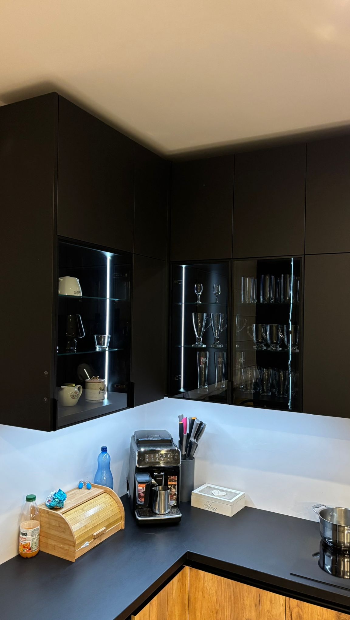 Nowoczesna, czarna kuchnia z podświetlanymi szklanymi witrynami, drewnianymi frontami i czarnym blatem. Na blacie ekspres do kawy i akcesoria kuchenne.