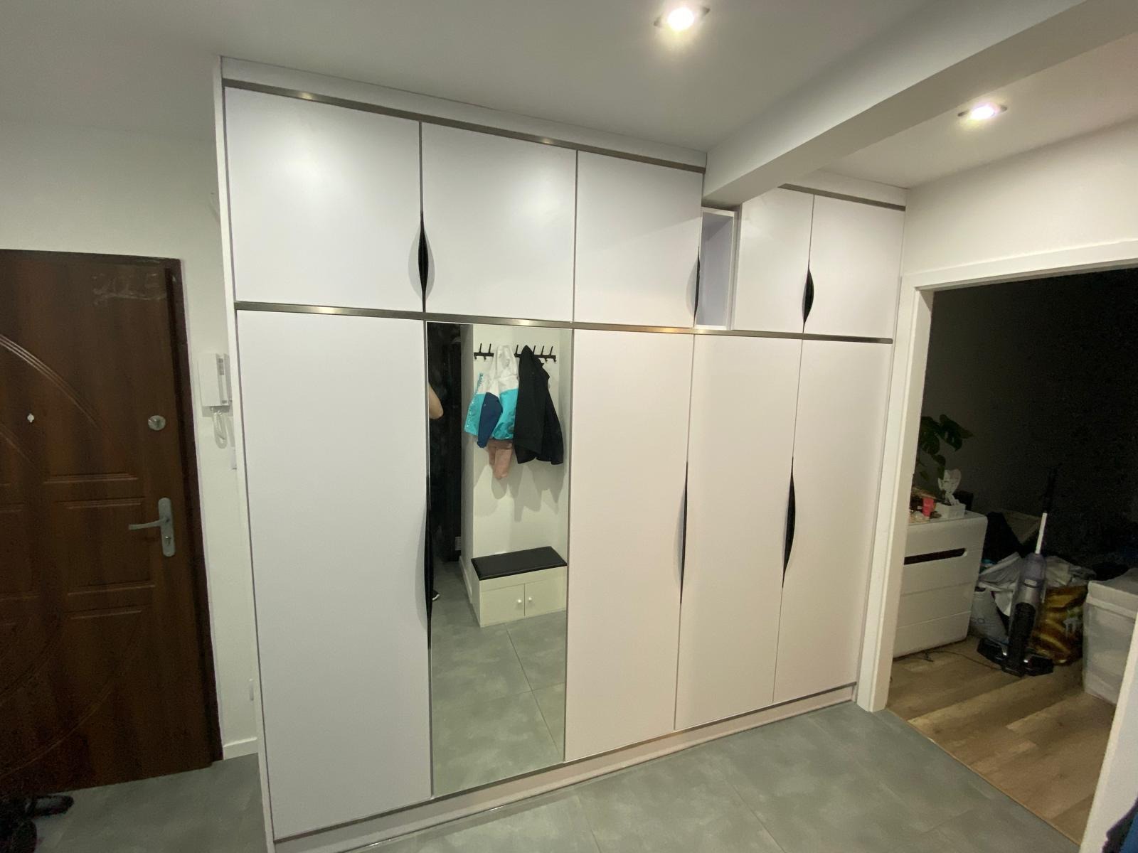 Biała szafa wnękowa z lustrem i minimalistycznymi uchwytami w przedpokoju. Widoczne ubrania wewnątrz szafy. Szara podłoga i drzwi wejściowe po lewej stronie.